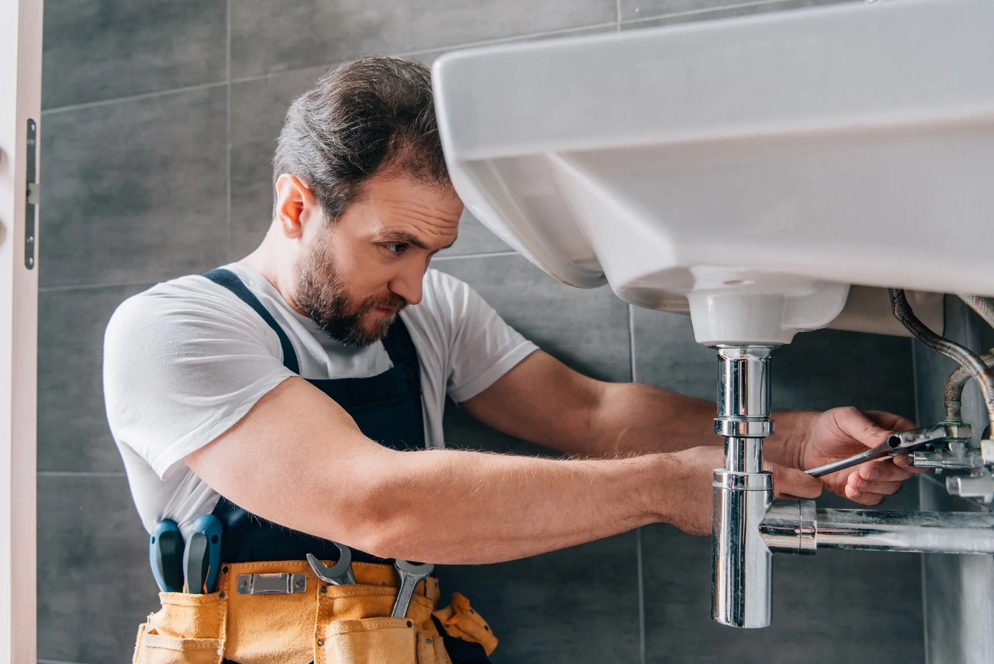 A man with a beard and gray hair is fixing a sink under a bathroom sink, wearing a tool belt with various tools.