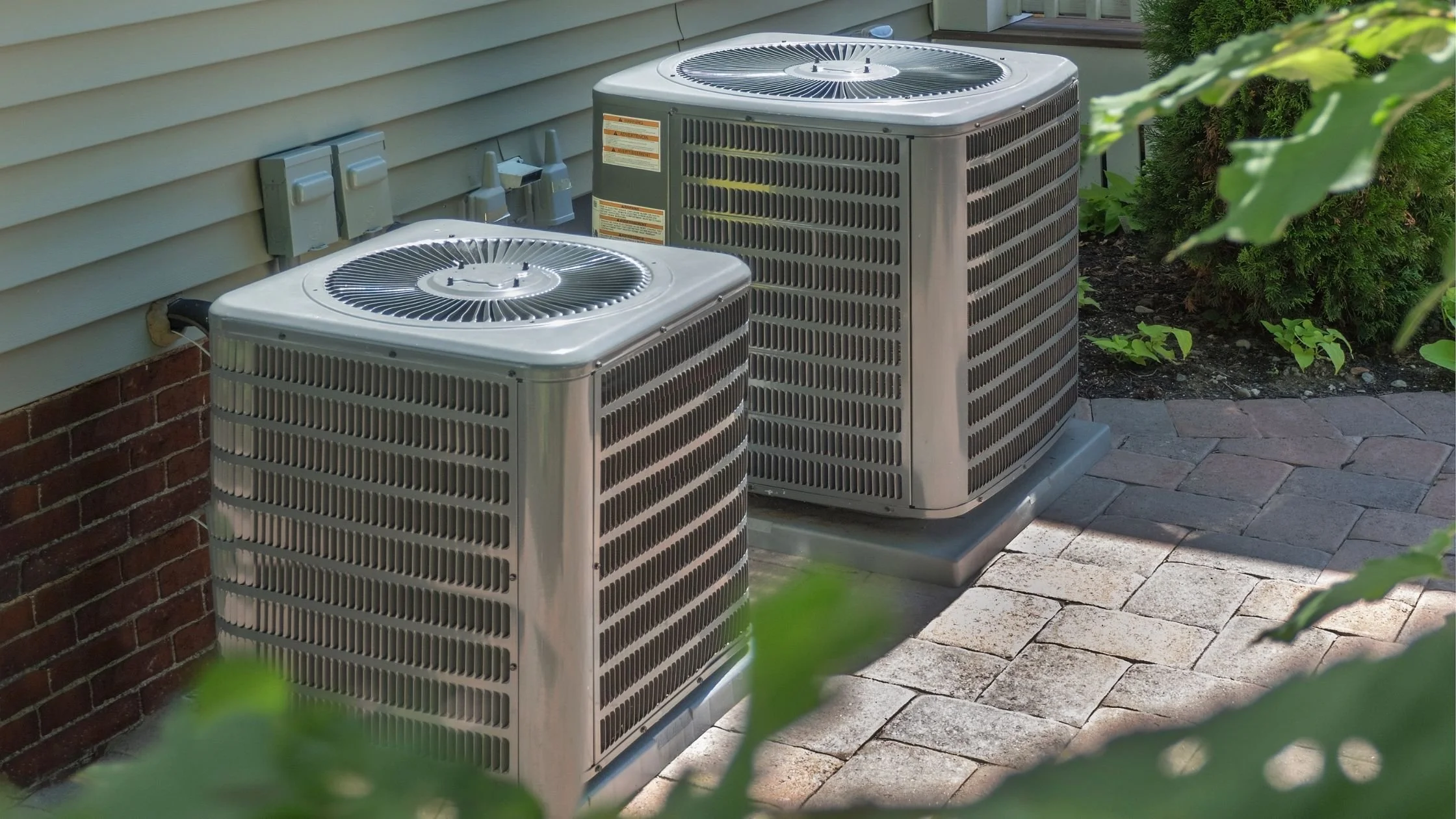 Two central air conditioning units outside a house on a stone patio, with siding and shrubbery nearby.