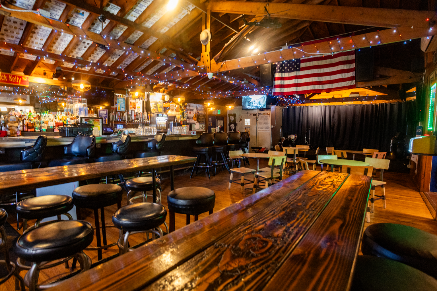 Interior of the "Club 416" bar with wooden decor, string lights, a long bar counter with bar stools, and various tables and chairs. An American flag hangs from the ceiling, and there is a black curtain in the corner.