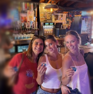 Three young women smiling with drinks at a bar, with a colorful background and bar equipment visible.