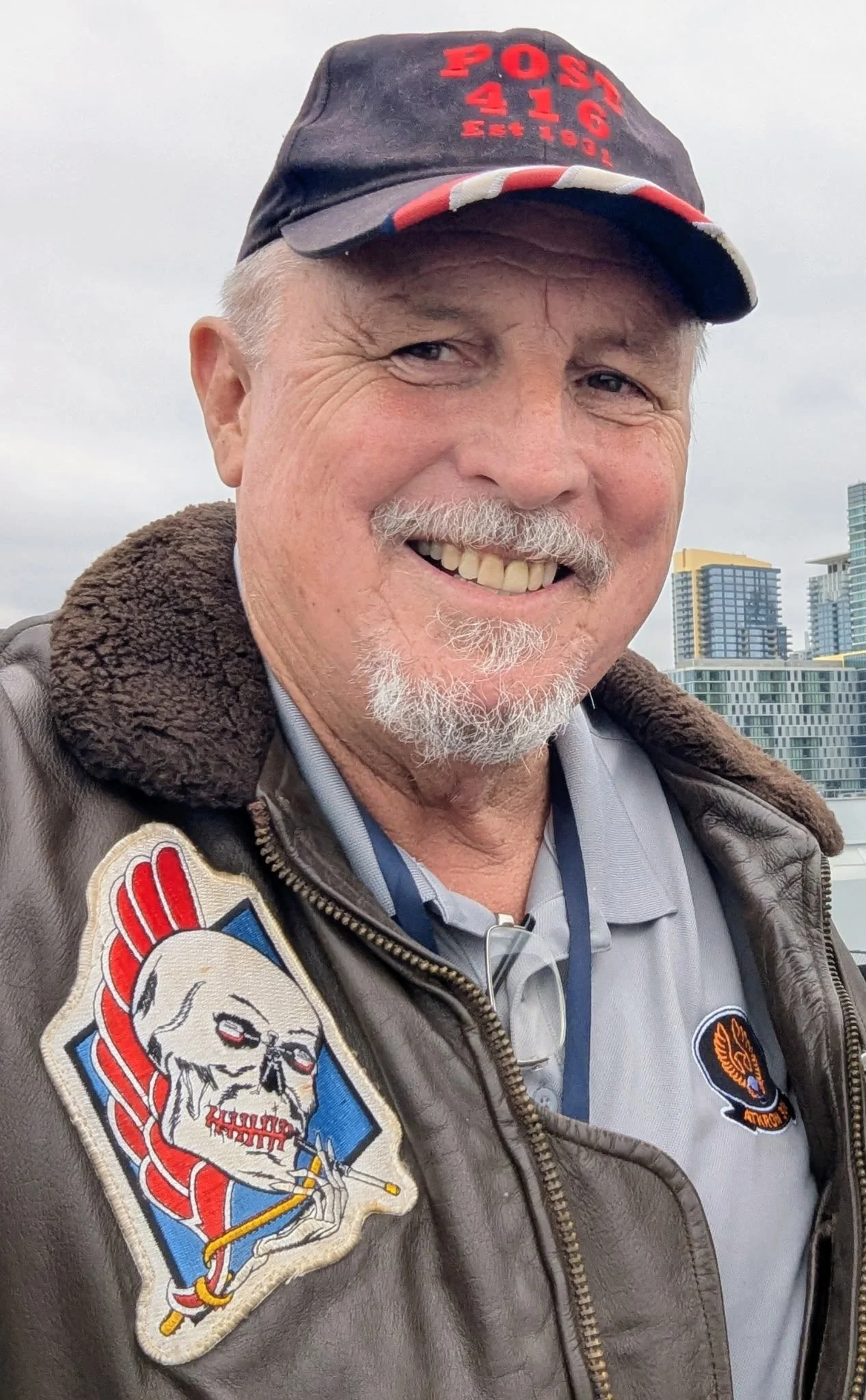 A smiling older man wearing a dark cap with red lettering, a brown leather jacket with patches, and glasses hanging from his collar, with a city skyline in the background.