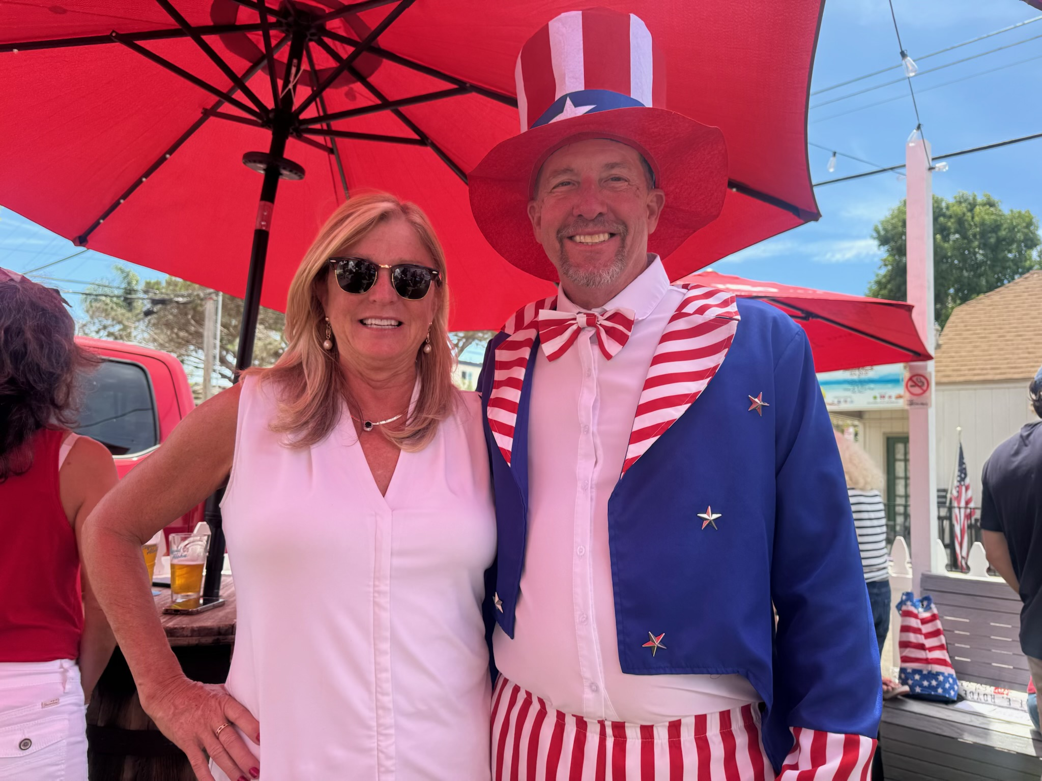A woman and a man dressed in patriotic American costumes standing under a large red umbrella. The man is wearing a red, white, and blue Uncle Sam outfit with a matching hat and bow tie. The woman is wearing a white sleeveless top and sunglasses. They are smiling at the camera, with other people, a drink, and a SIDEBAR sign visible in the background.