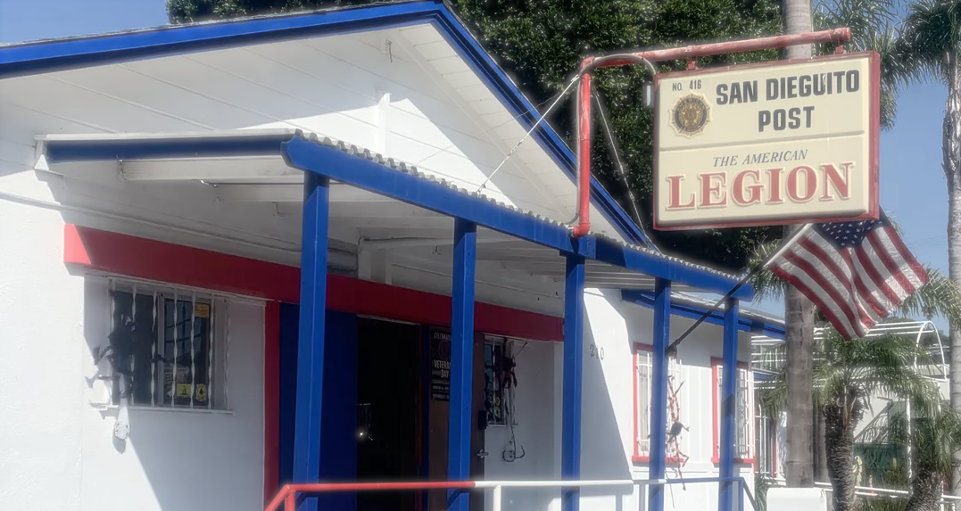 The San Dieguito Post, a building with a blue and white exterior, features a red border above the entrance, a sign indicating it is the American Legion San Dieguito Post 416, and an American flag flying nearby.