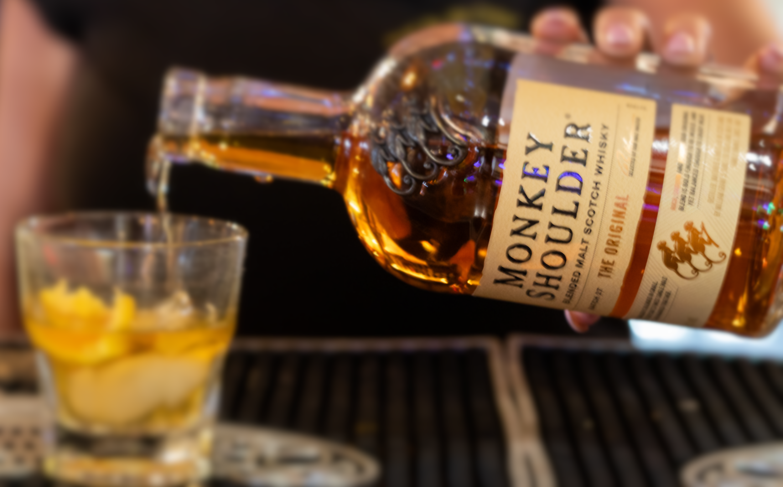 A hand holding a bottle of Monkey Shoulder Scotch whisky, with a glass of whisky with ice and lemon slices in the foreground.