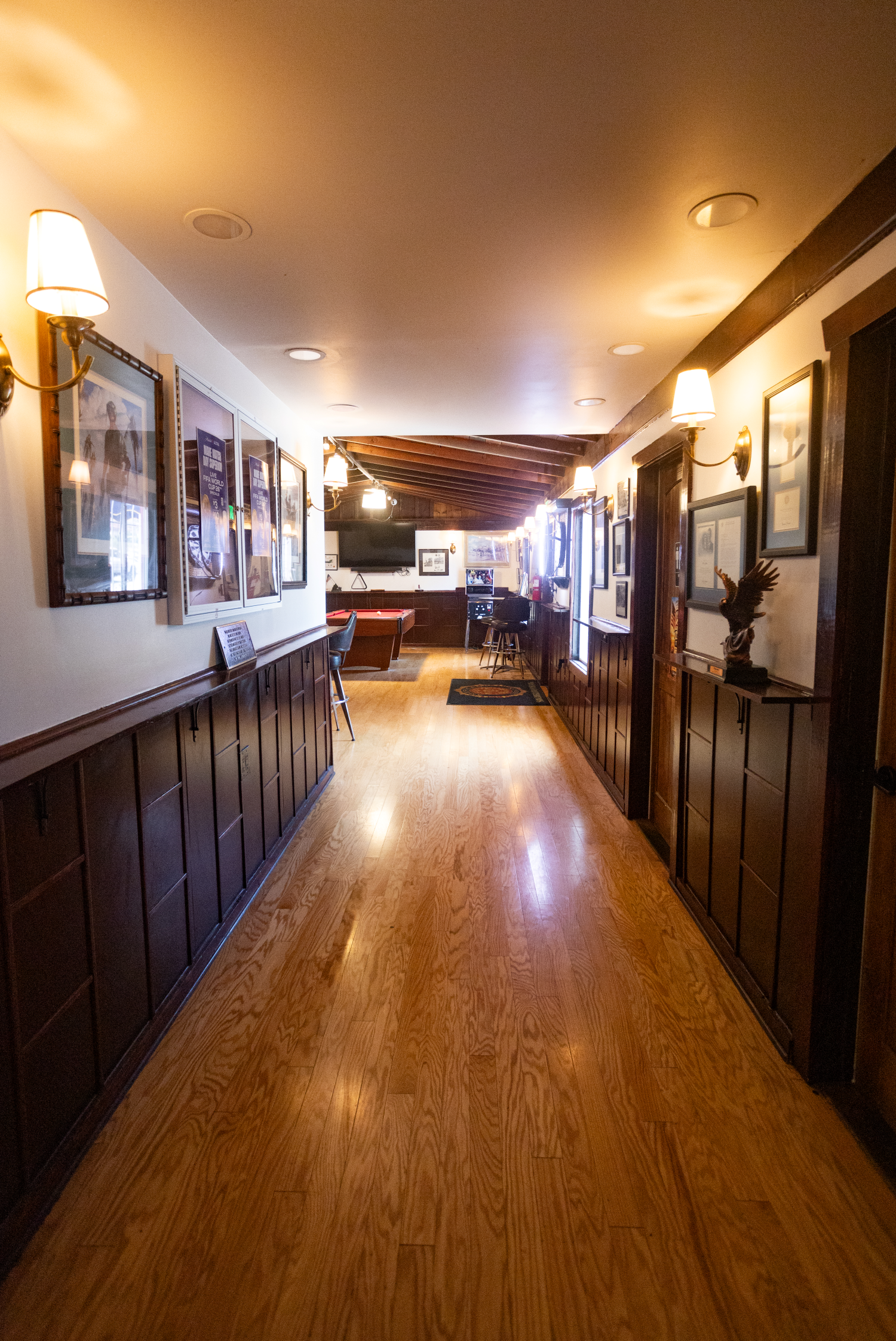 Long hallway of the American Legion San Dieguito Post 416 with hardwood flooring, framed veteran-centric pictures on white walls, wood paneling, wall-mounted lamps, and a pool table at the far end.