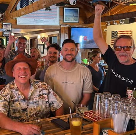 Group of people enjoying drinks at a bar, smiling, some raising their hands, with a television and American flag in the background.
