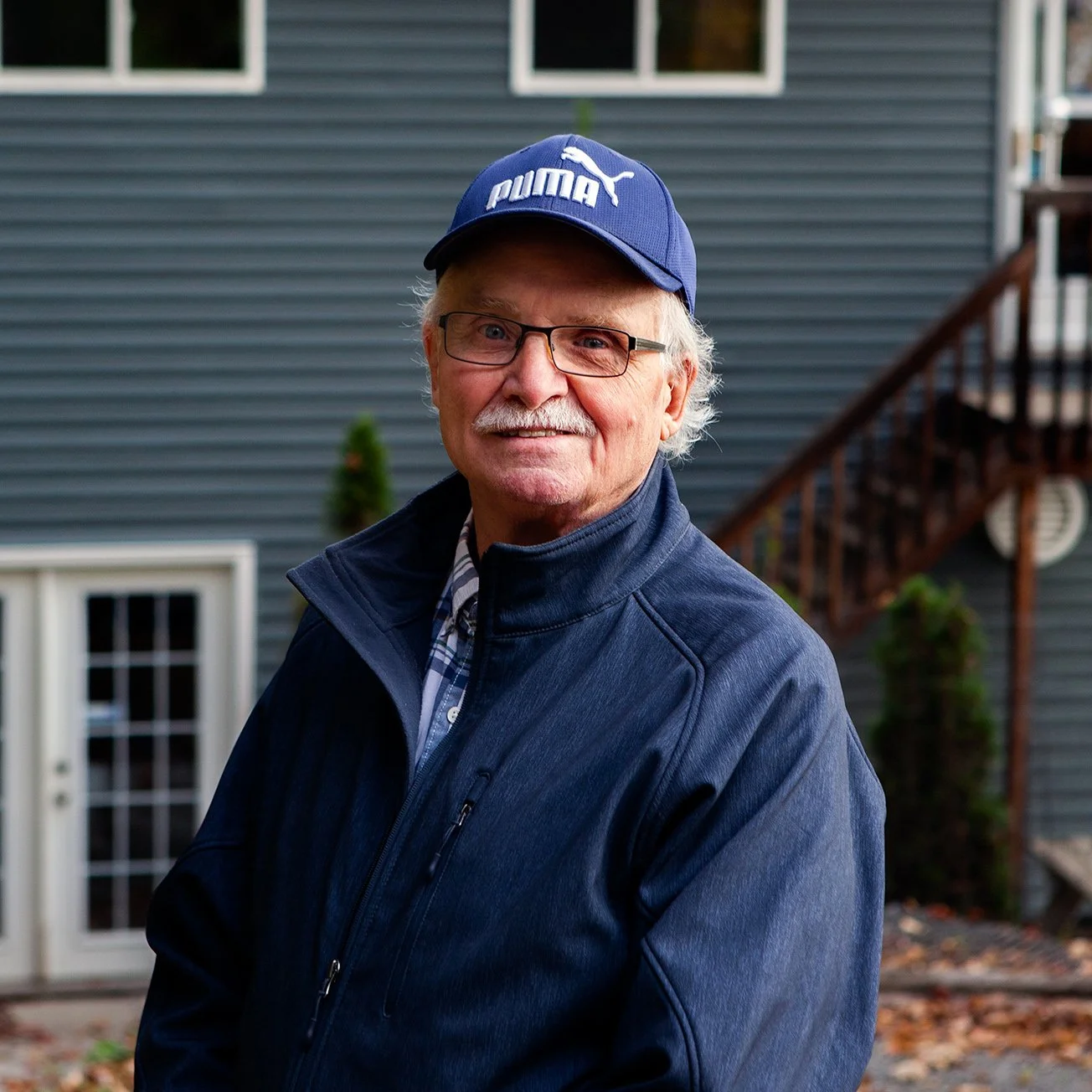 A picture of the founder and owner of True view Design with gray hair and glasses, wearing a blue Puma cap and a navy blue jacket, standing outside in front of a boathouse with gray siding and a staircase, smiling.