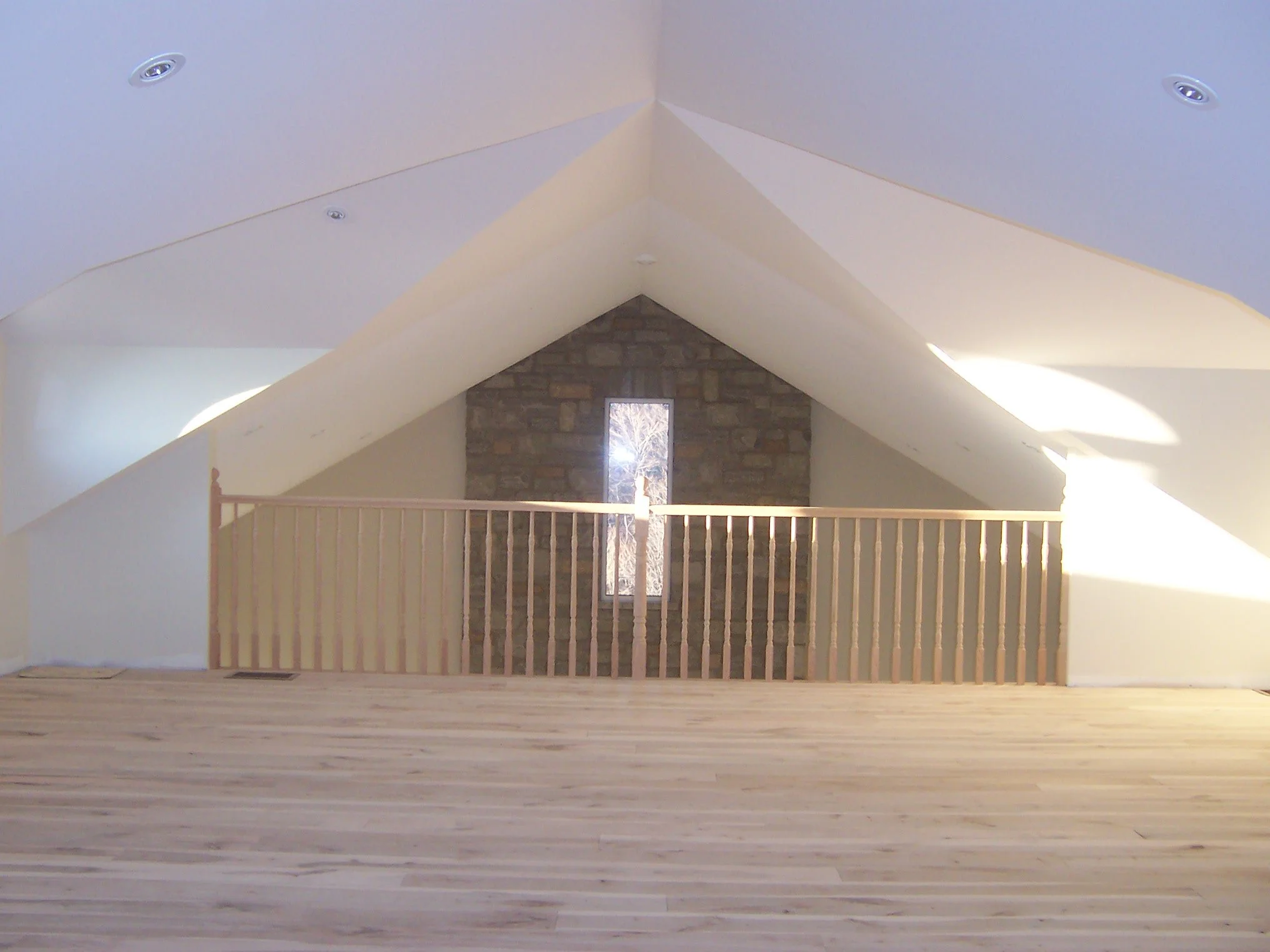 Interior view of a room with a vaulted ceiling overlooking another lower level room showing a stone fireplace chimney with a window inset in the stone,  light-colored walls, and a wooden railing in the foreground.