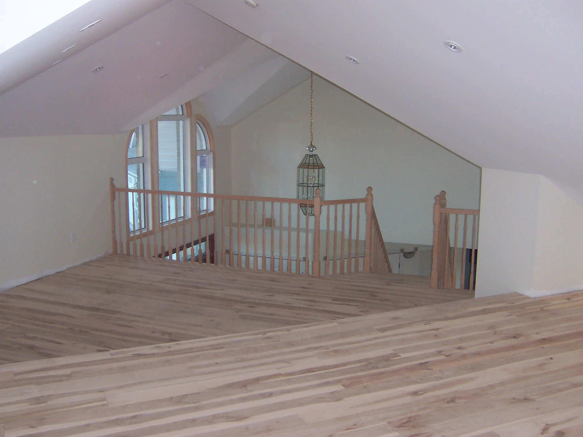 An upper loft area with wood flooring and wood railing that overlooks a grand front entry with arched windows and a hanging chandelier.