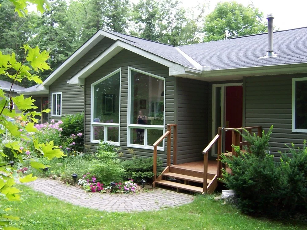A house with textured green siding, a gable roof, and a small porch with wooden stairs, surrounded by green plants and colorful flowers.