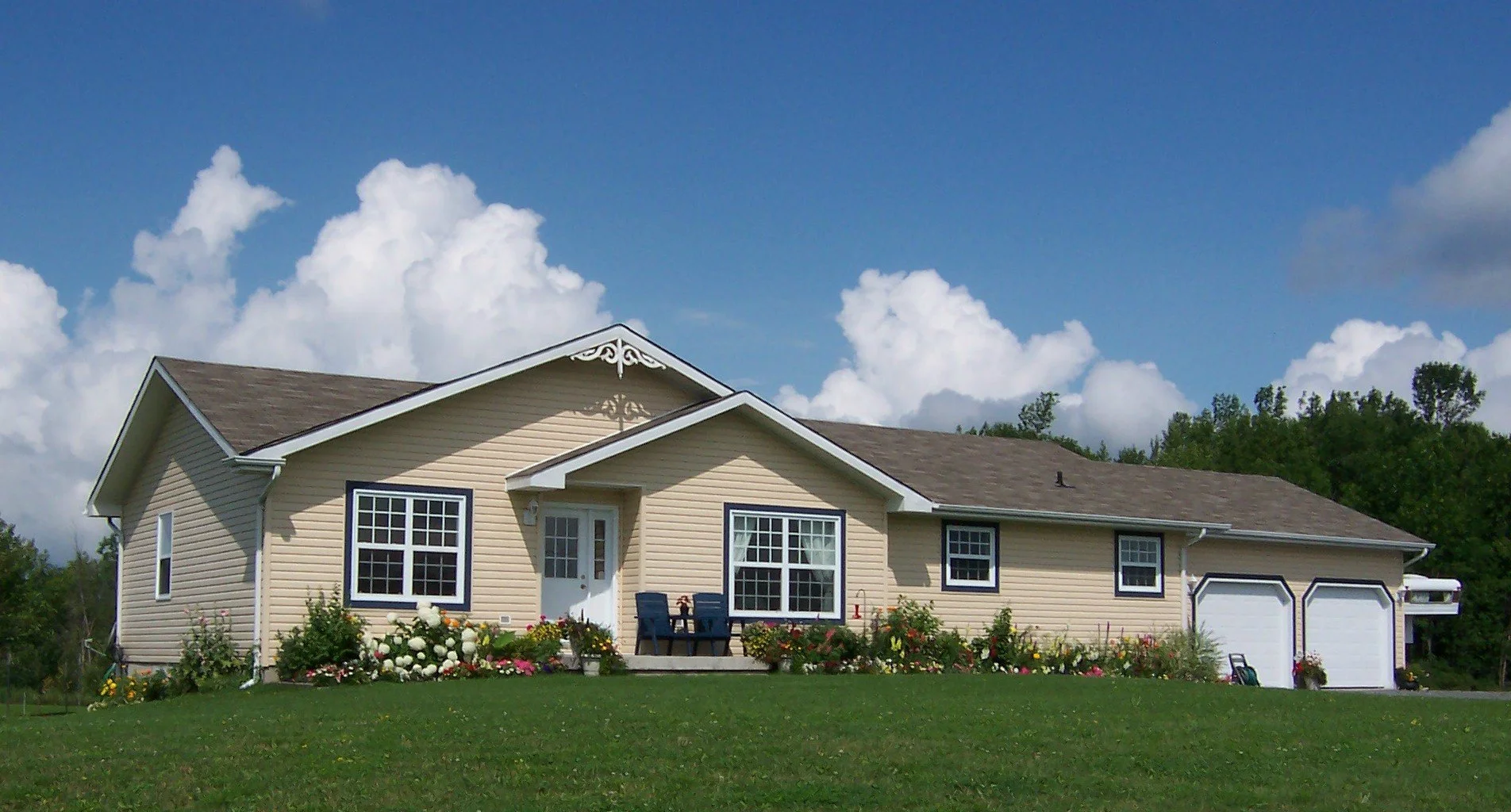 A pleasant yellow sided , bungalow with a front porch, surrounded by a garden with colorful flowers, under a partly cloudy sky with some trees in the background.