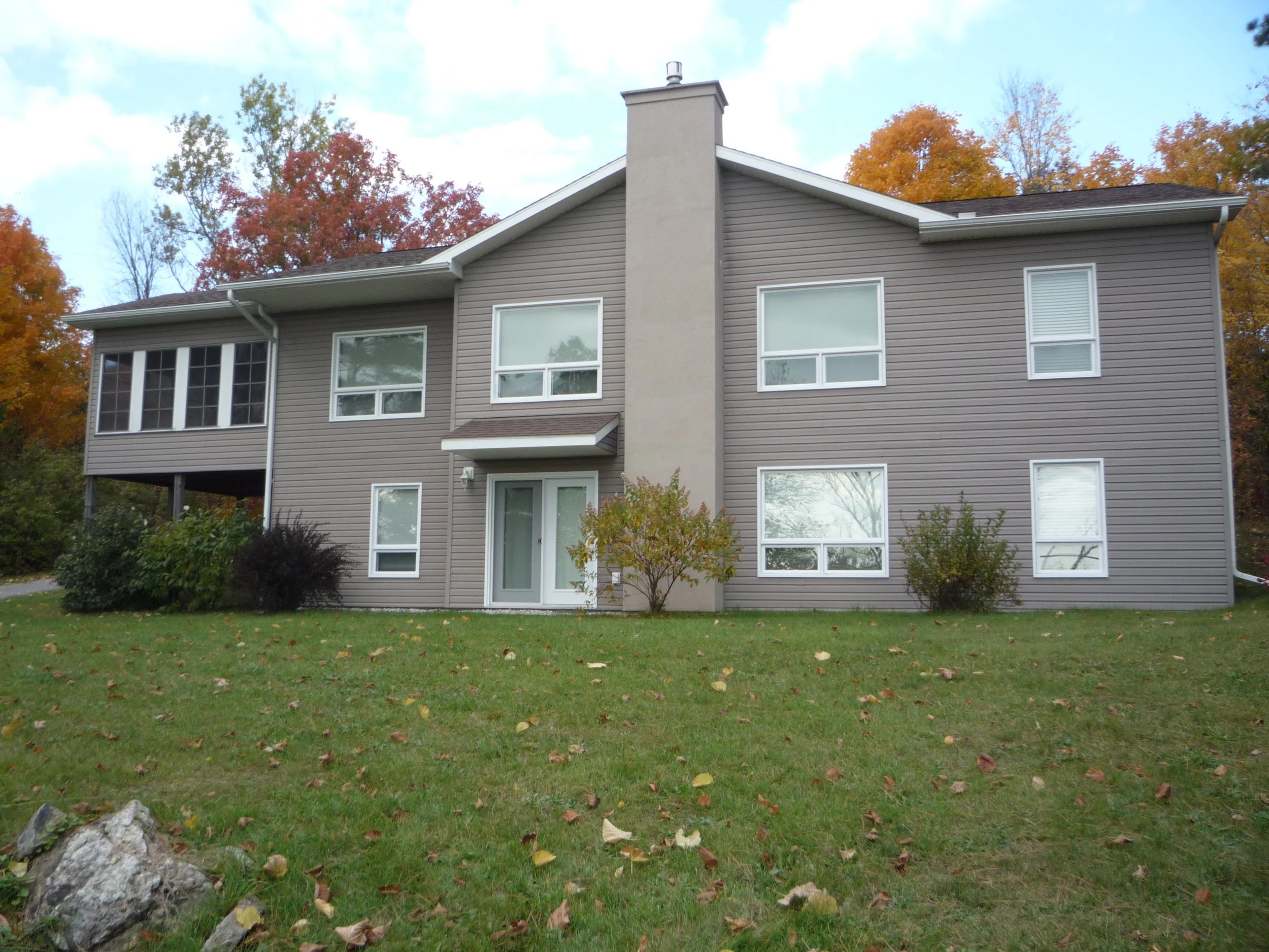 The back of a gray, multi-story residential building with several windows and a central chimney, surrounded by fall foliage and a green lawn with fallen leaves.