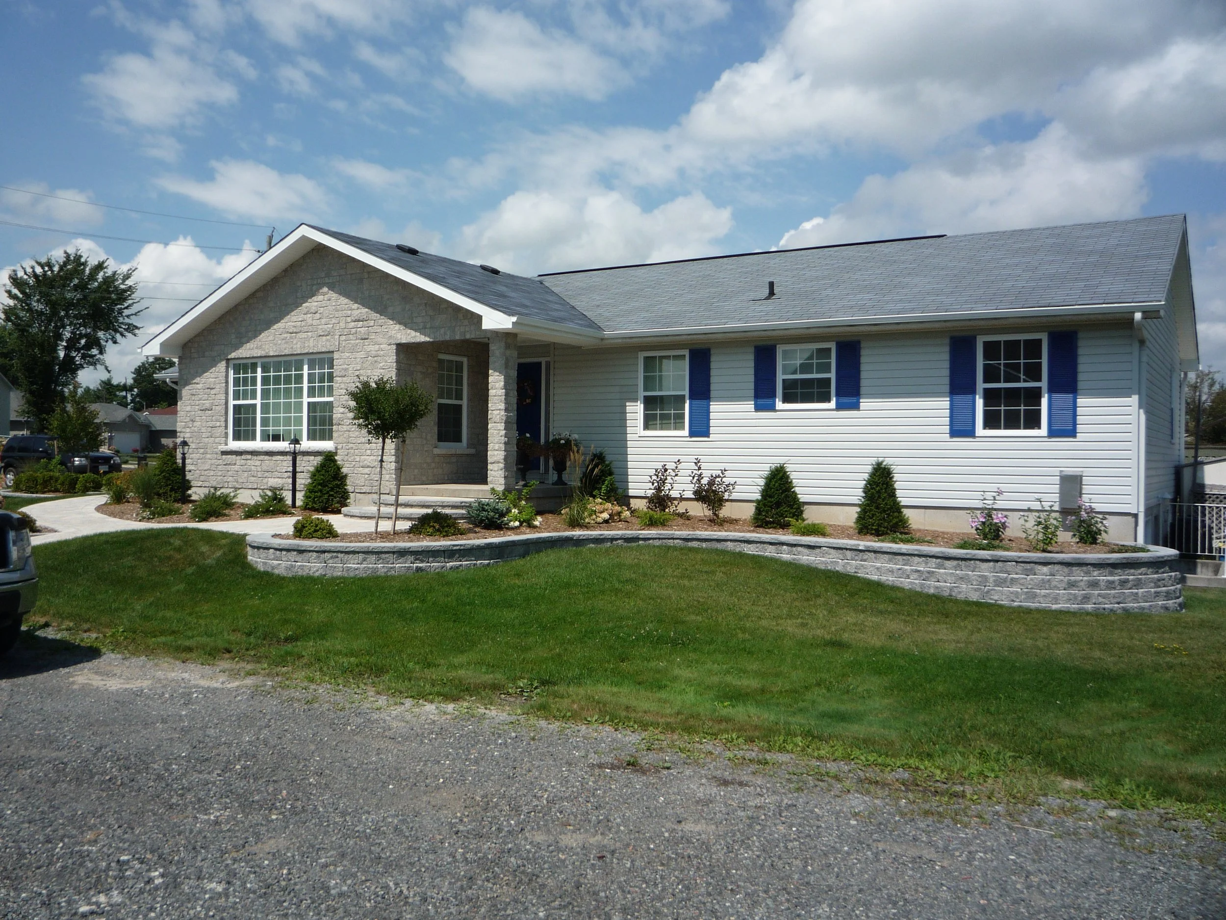 A suburban house with a stone and siding exterior, a blue shingle roof, and blue shutters, with a manicured front yard, a curved walkway, and a gravel driveway on a sunny day with a partly cloudy sky.