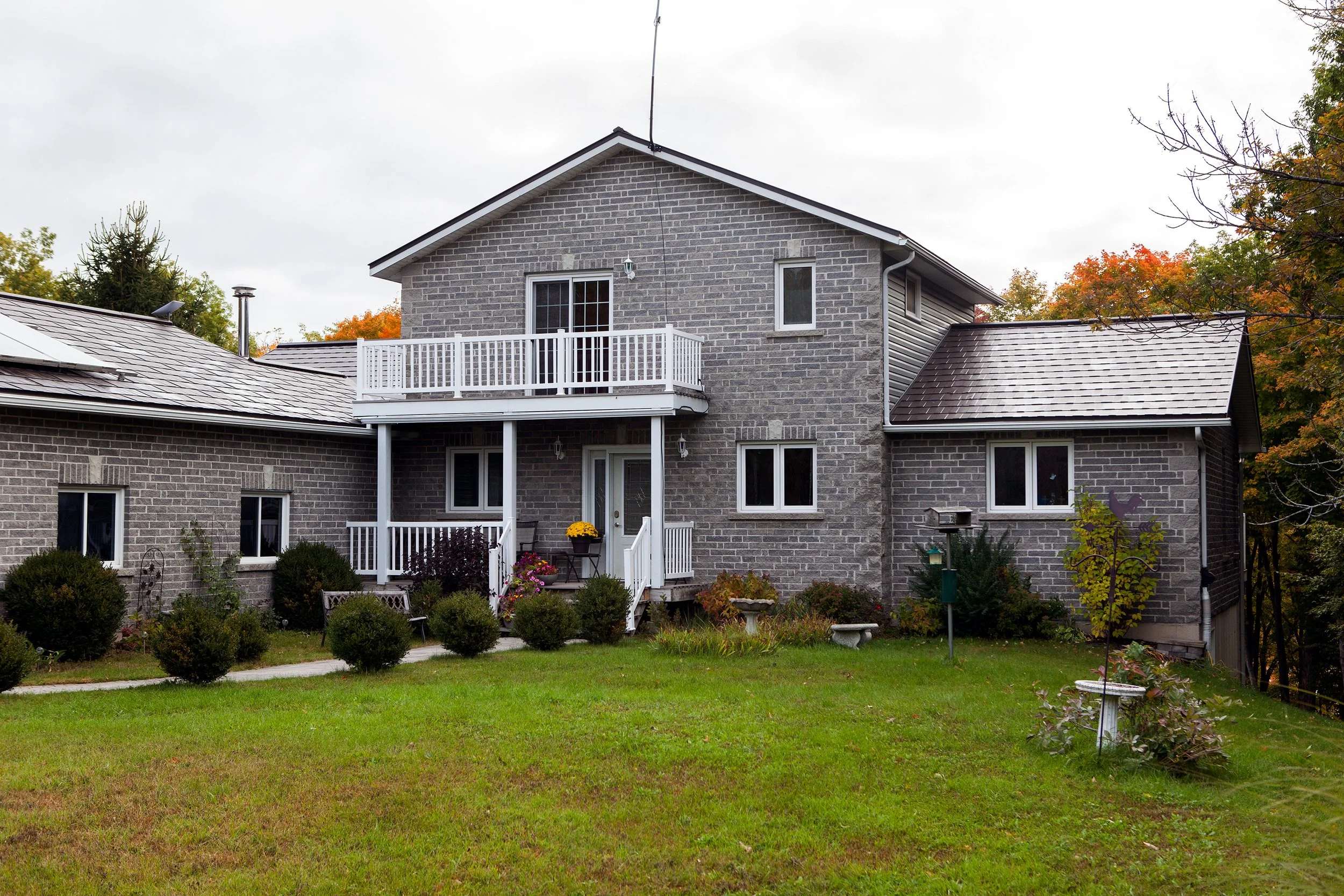 A gray brick house with a small white balcony and a garden in the front yard, autumn trees in the background, and an overcast sky.
