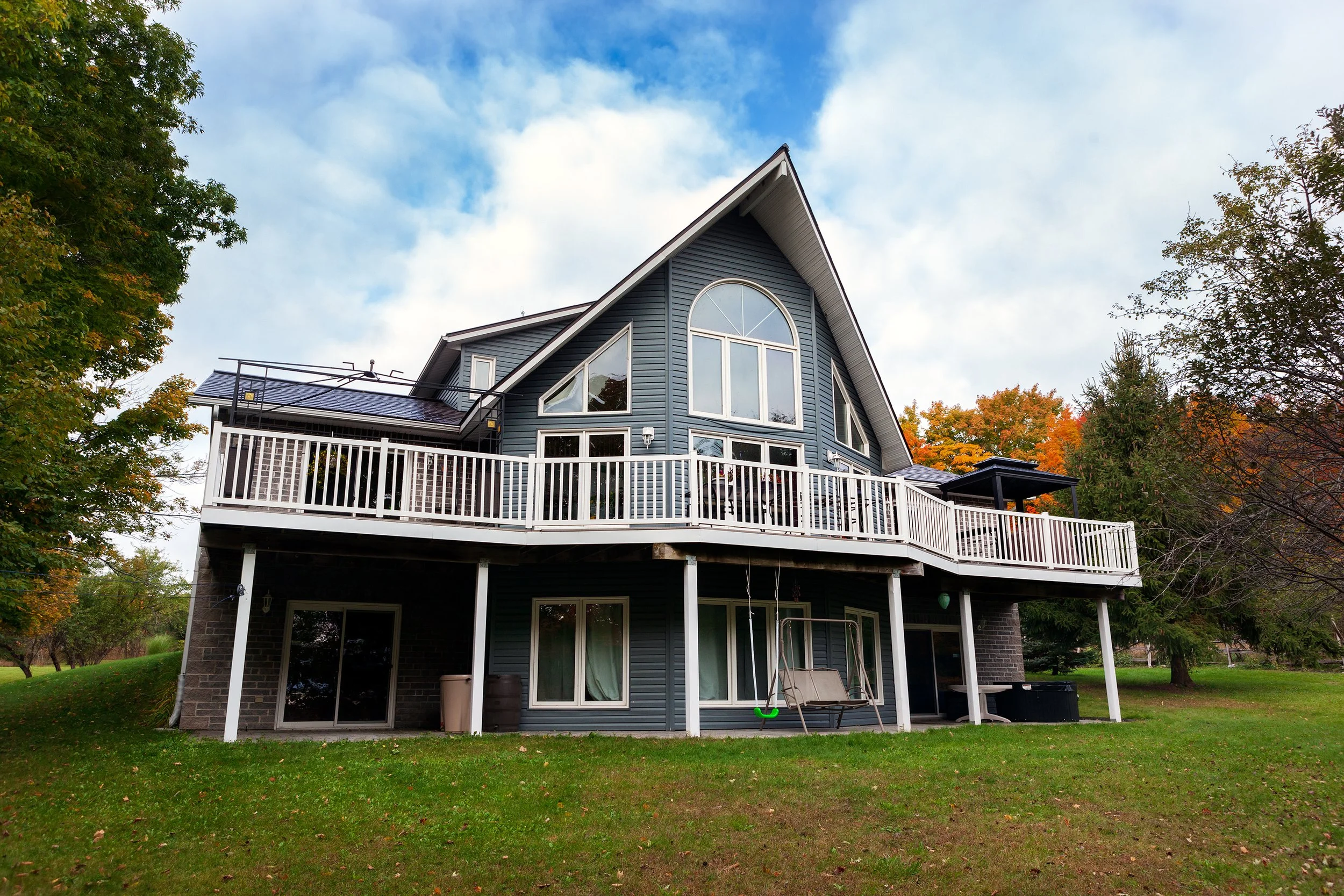 A blue sided house with walk out foundation level, large deck , white deck railing, architectural shaped windows, surrounded by trees with autumn foliage, under a cloudy sky.