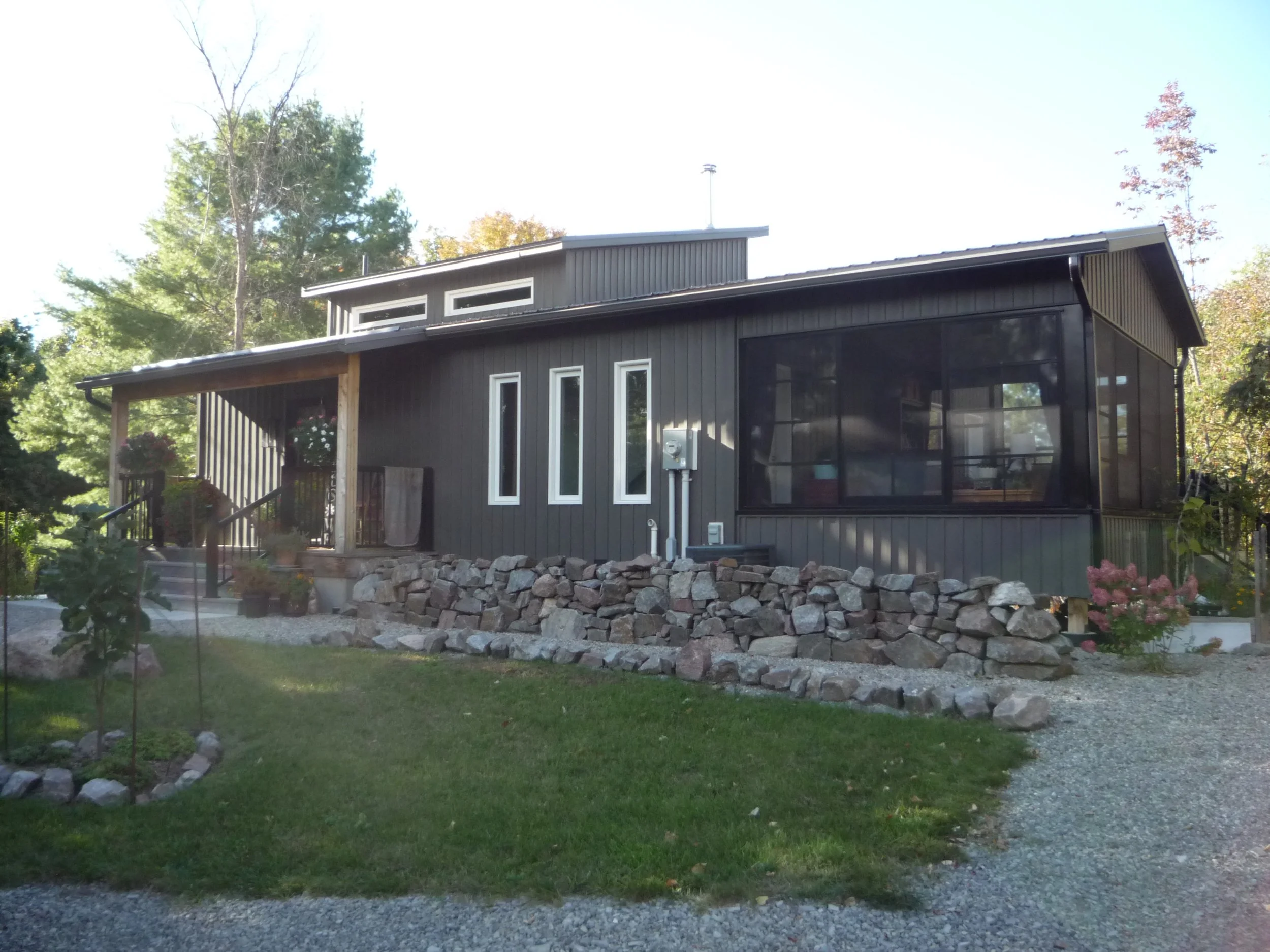 A modern black house with a sloped roof and tall, narrow windows, surrounded by trees and a landscaped yard with a rock wall and green grass.