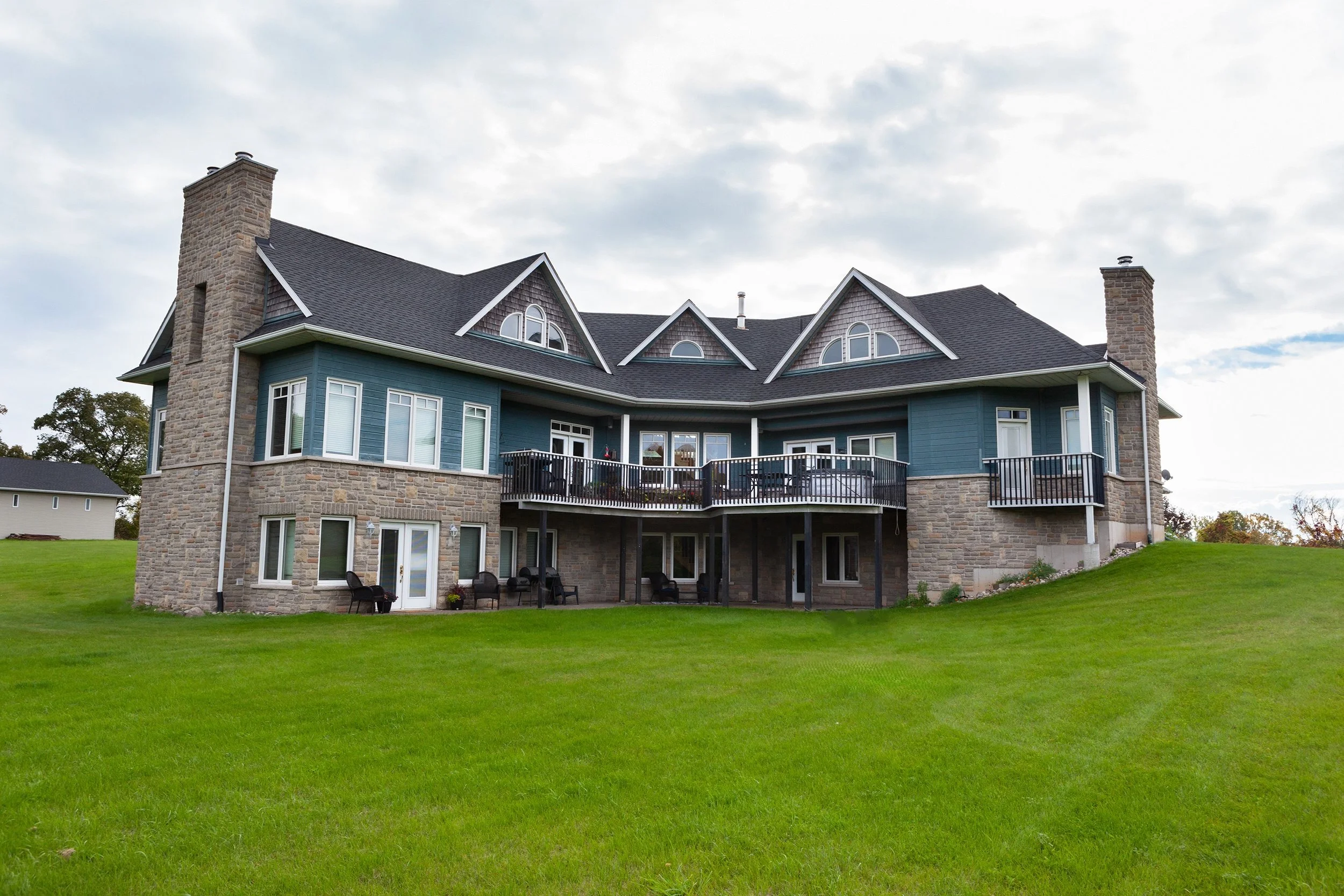 Large house with blue siding and stone accents, multiple gabled roofs, and chimneys, set on a well-maintained grassy yard during daytime with a cloudy sky.
