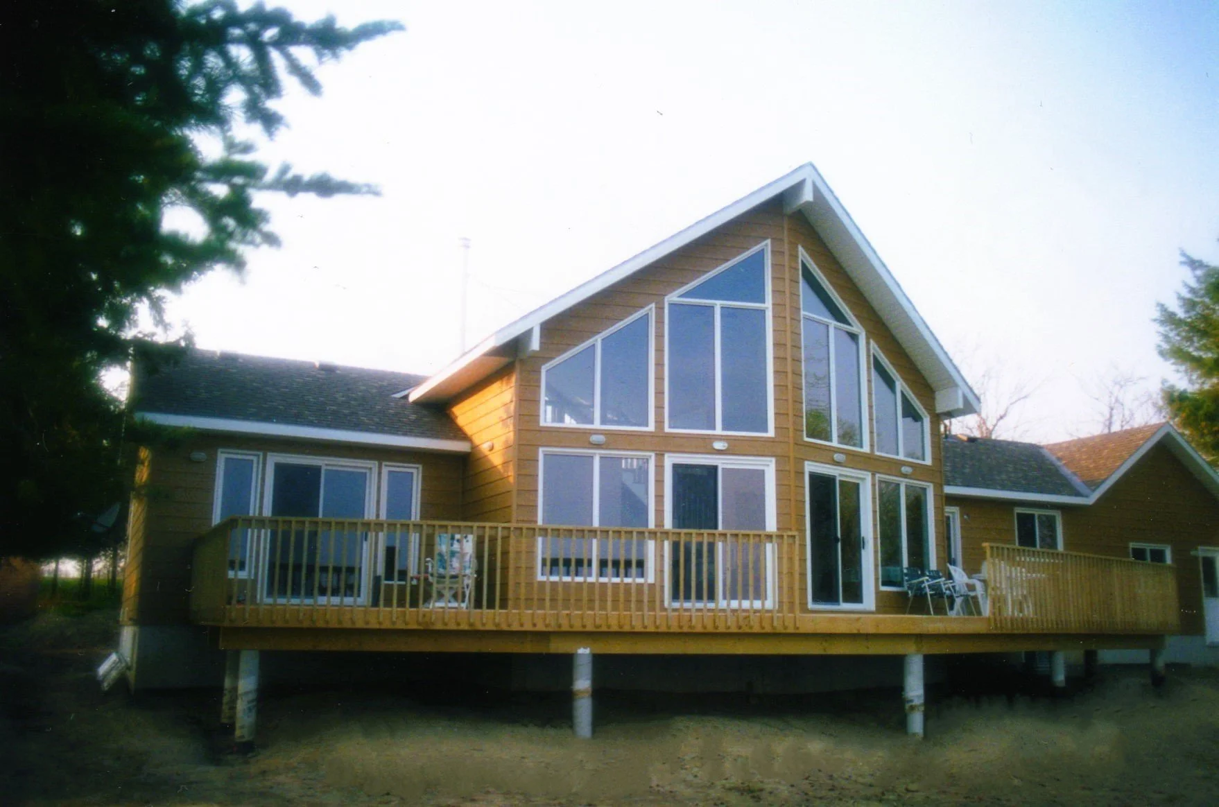 A two-story house with large triangular and rectangular windows, a wooden exterior, and a deck with chairs, elevated above the ground with support pilings. Trees are visible around and behind the house.