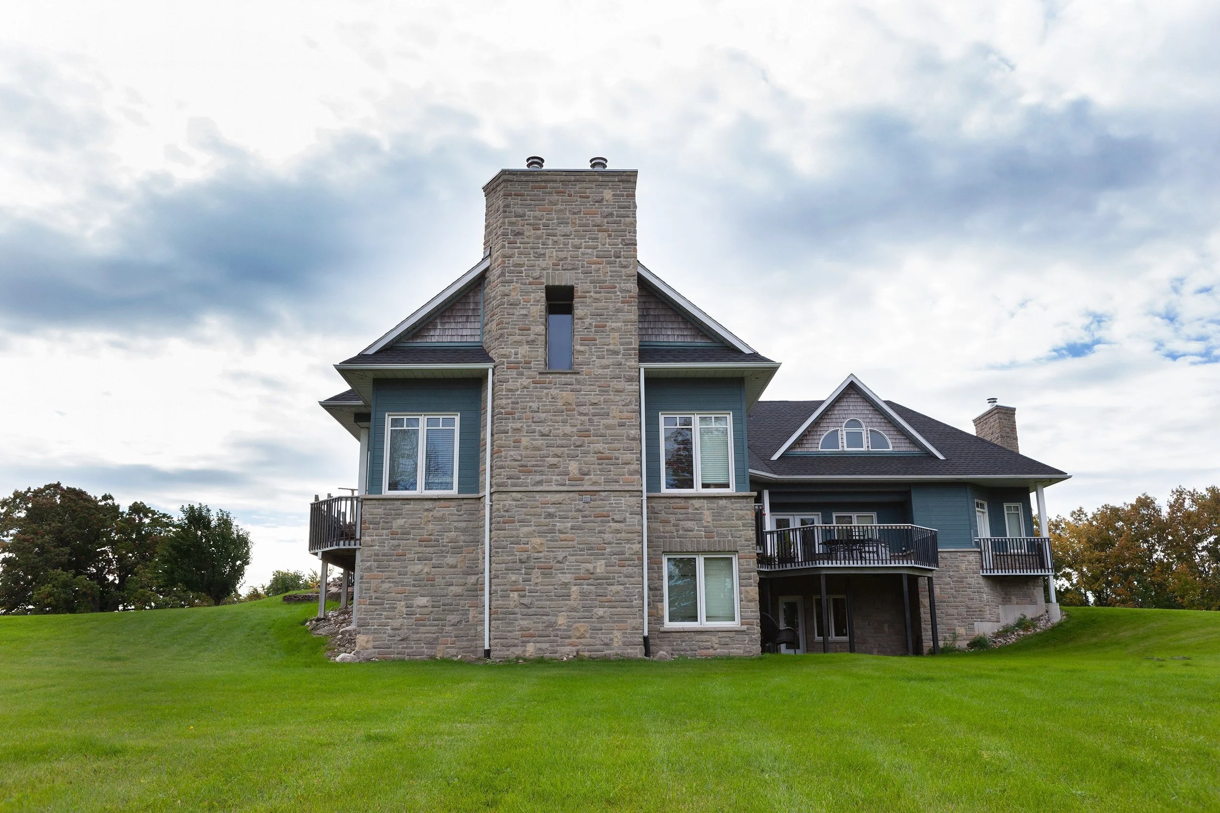 A large house with a stone chimney, blue siding, multiple windows, and a backyard deck, set on a grassy lawn with trees in the background under a cloudy sky.