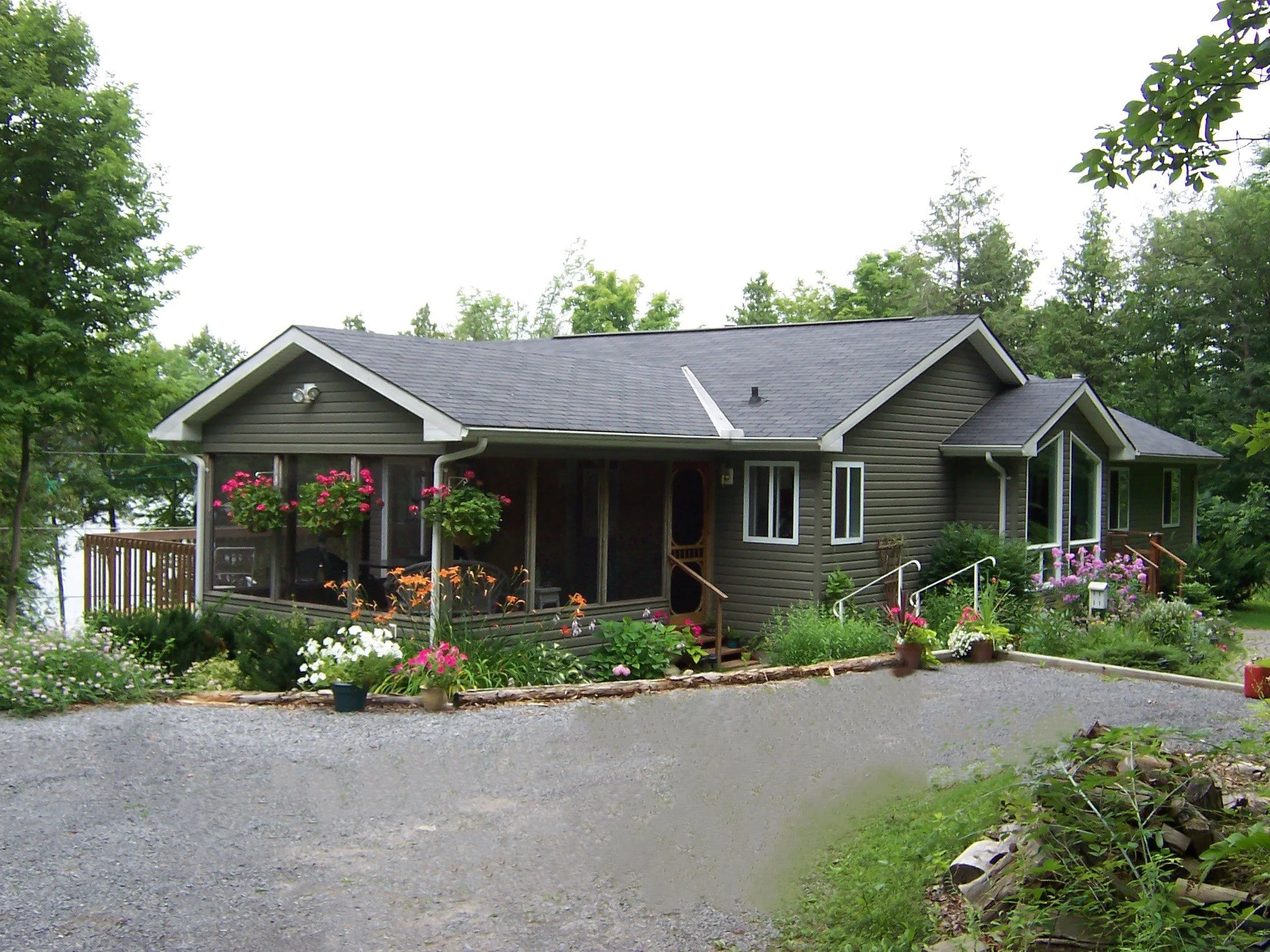 A green sided  country house with a gabled roof, attached screen roomsurrounded by a lush green garden, featuring colorful flowers and potted plants, with a gravel driveway in the foreground.