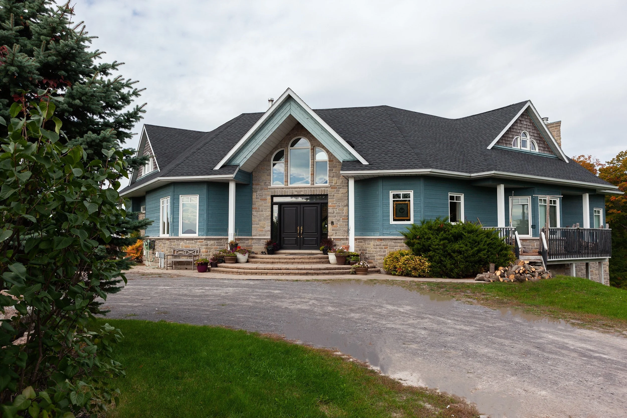 Front view of a large custom house with a stone and blue siding exterior, gabled roof, and multiple windows, surrounded by greenery and a gravel driveway.