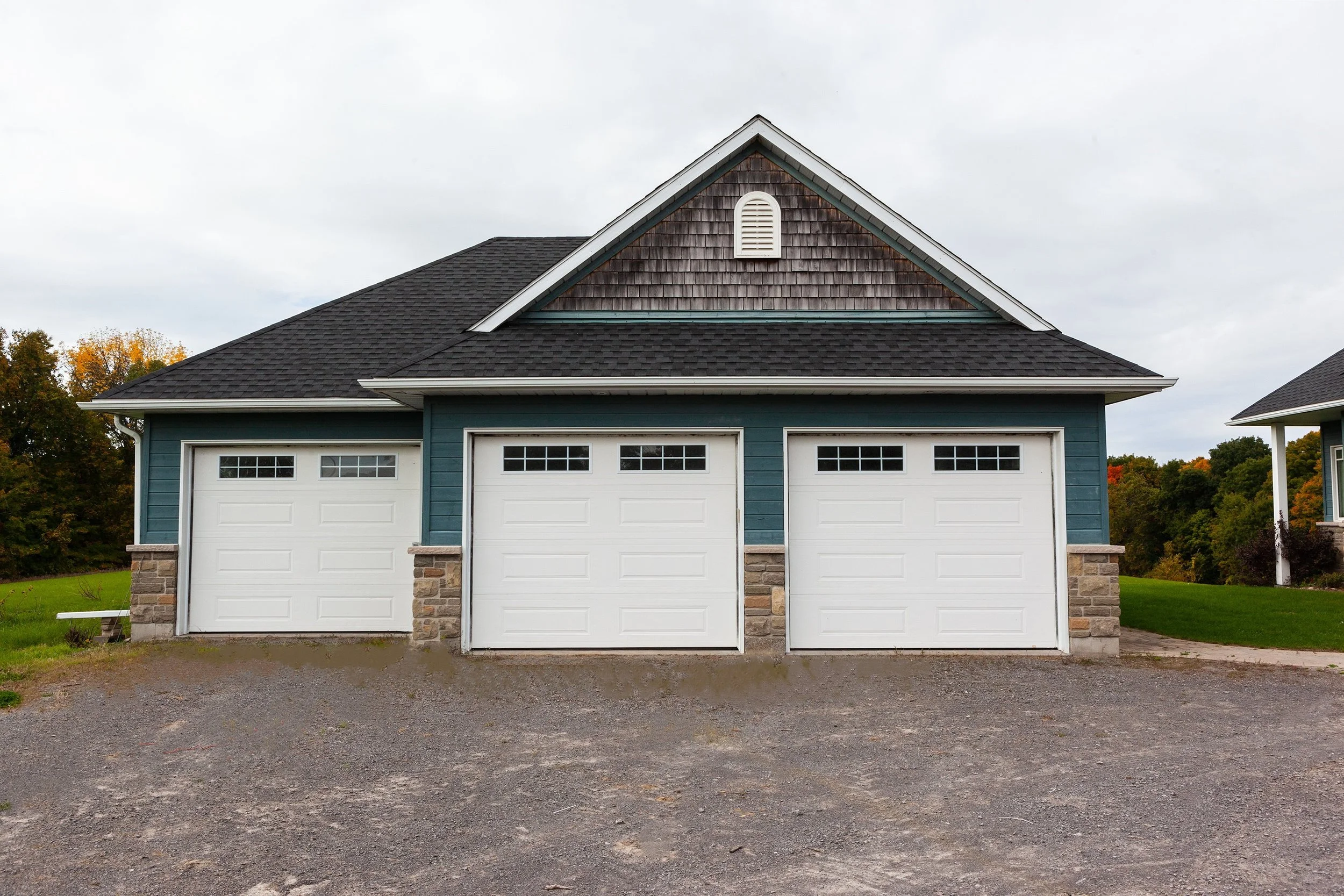 A three-car garage with a gable roof, blue siding, stone accents at the base, and a vent on the front gable, with trees in autumn foliage in the background and a gravel driveway in front.