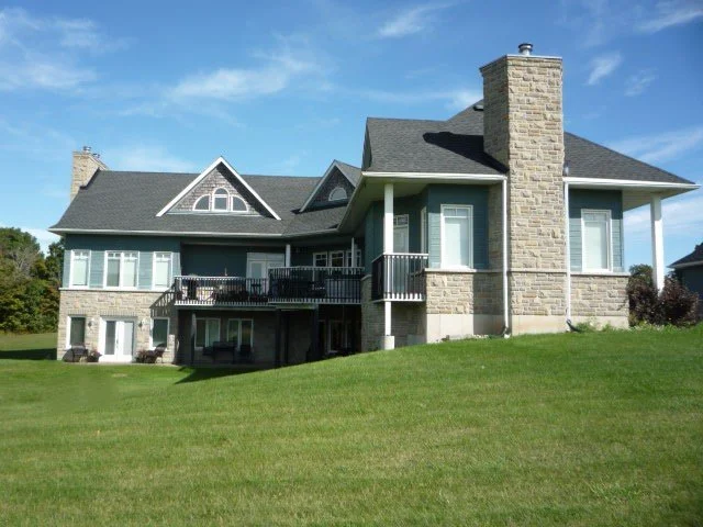 Large house with stone chimney, multiple gabled roofs, sundeck and a balcony, situated on a grassy lawn under a blue sky.