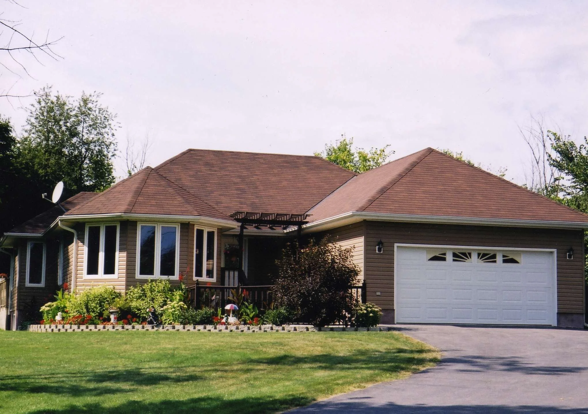 A mid brown color sided house with a brown pitched roof, garage door, bay windows, and white door with black handle, surrounded by a green lawn and a garden with plants and flowers. Trees are in the background and the sky is partly cloudy.