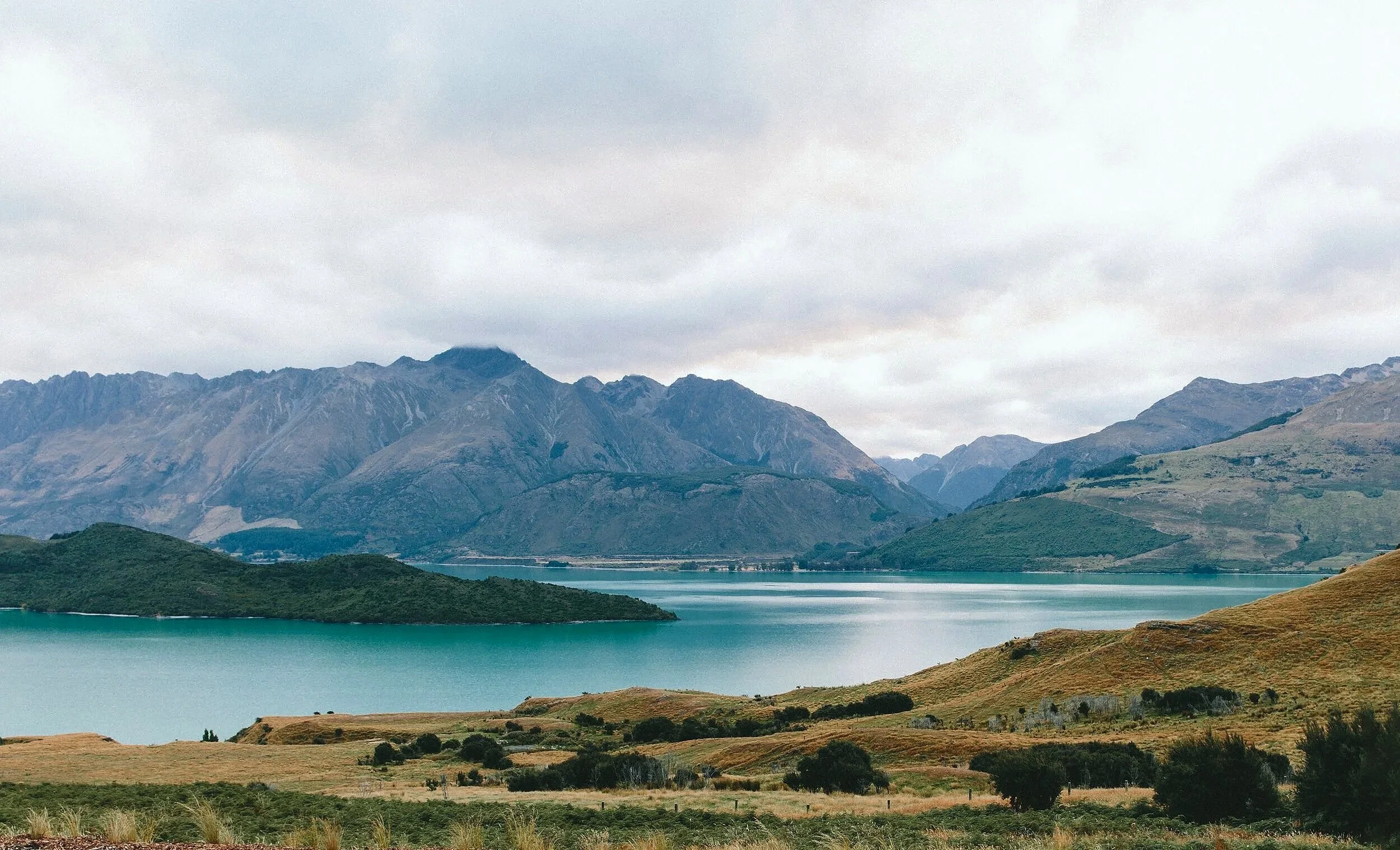 Scenic landscape with a large body of water, green hills, and mountain range under cloudy sky.