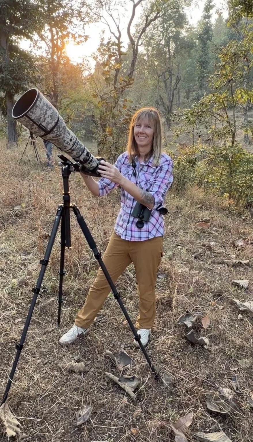 A woman with blonde hair in outdoor attire, standing in a wooded area, smiling and holding a large telescope on a tripod during sunset.