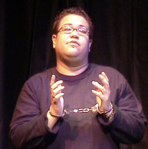 Person with short hair and glasses standing in front of black curtains, wearing a dark sweatshirt, with handcuffs on their wrists and their hands raised.