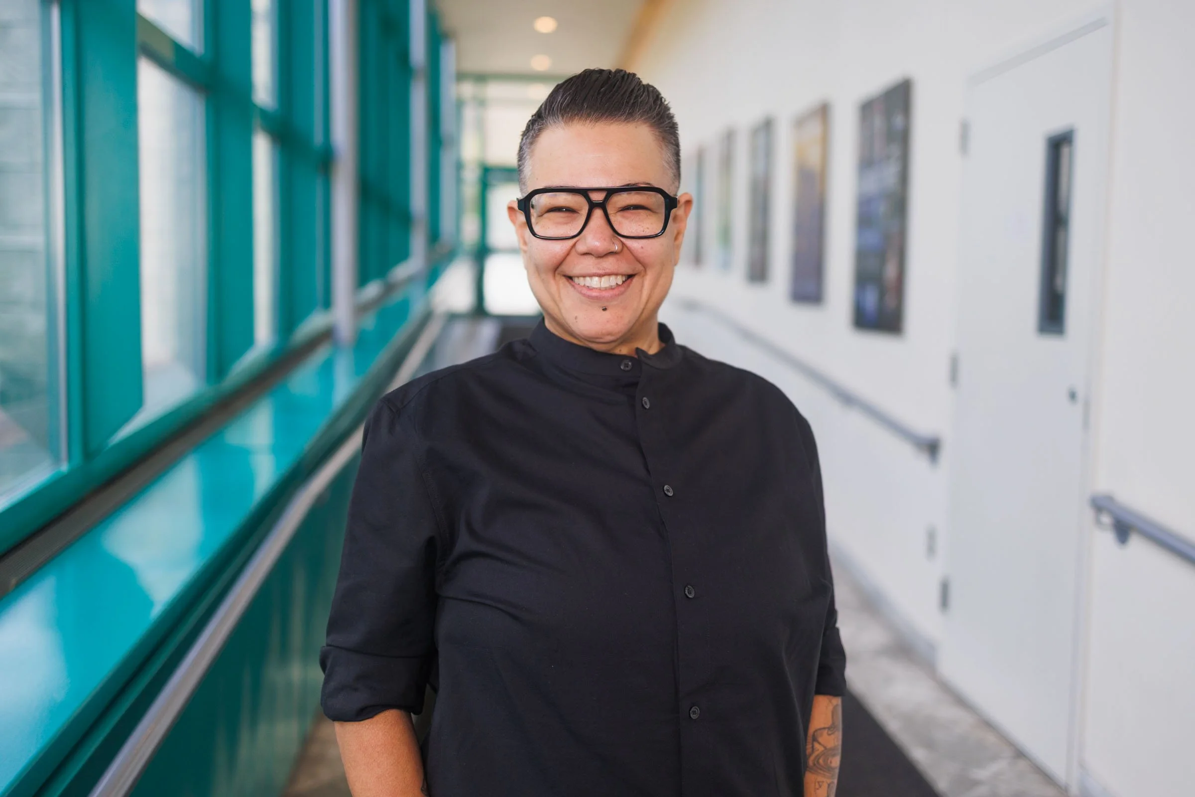 Smiling woman with glasses and tattoos standing in a brightly lit corridor with framed pictures on the wall.