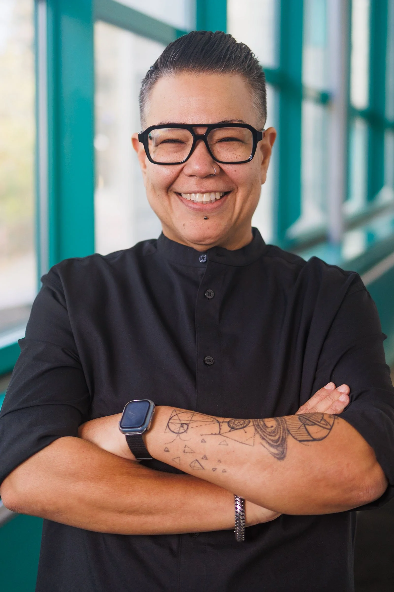 A person with short hair, glasses, and tattoos on their arm smiling with arms crossed, inside a building with large windows.