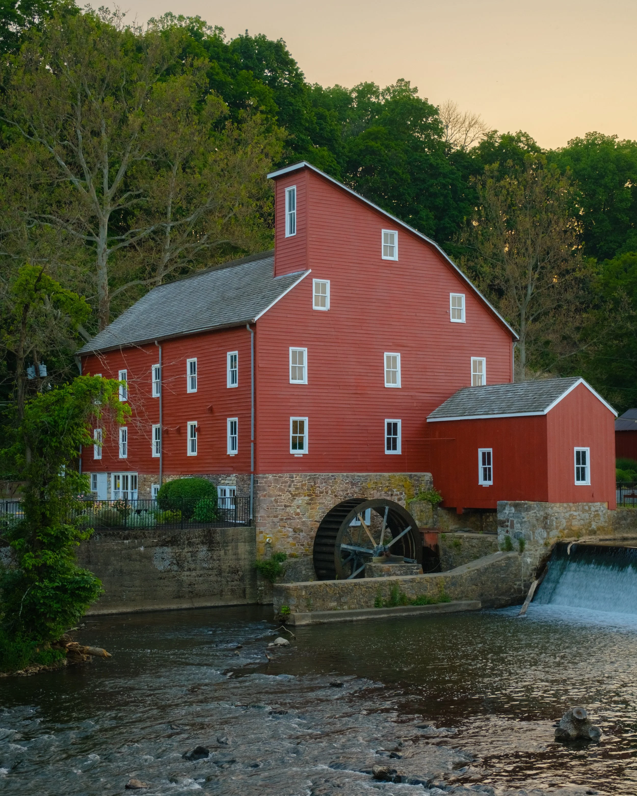 The historic Clinton Red Mill. A large red mill with white trim and multiple windows situated along a river, with a water wheel attached to its lower level.