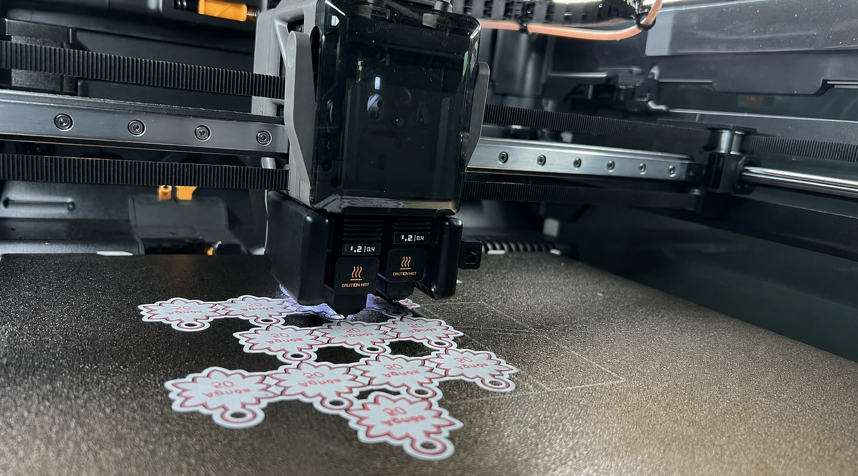 Une machine d'impression 3D en fonctionnement, déposant des autocollants en forme de nuage avec le texte '50ans' sur une surface noirâtre.