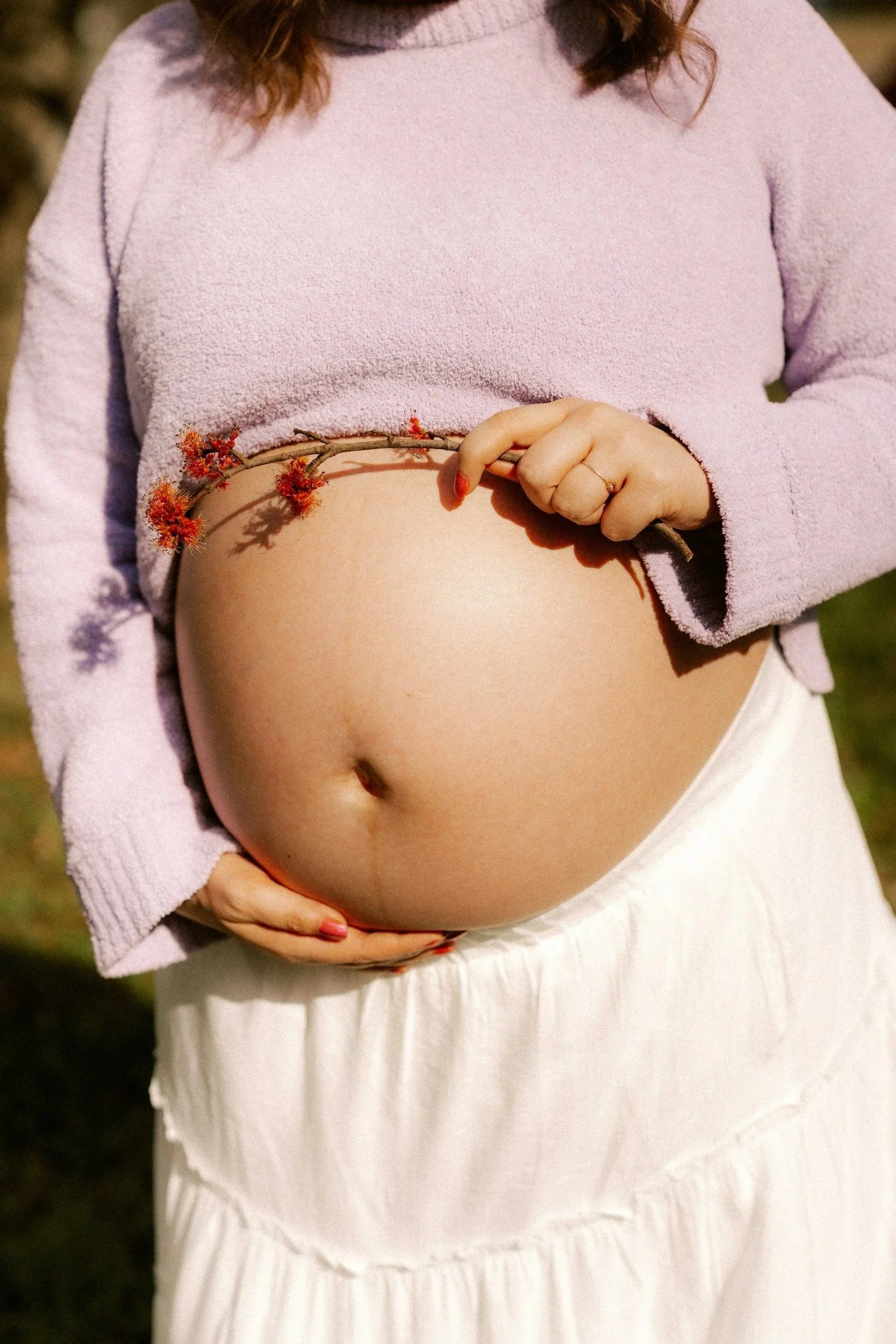 Eine schwangere Frau hält einen Ast mit roten Blüten vor ihren Bauch, trägt einen lila Pullover und einen weißen Rock