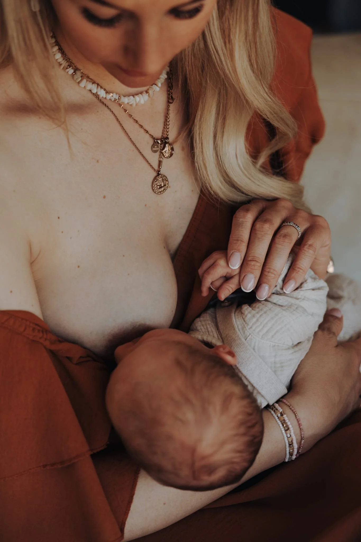 Eine Frau mit blonden Haaren, mehrere Halsketten und Ringen, die ein Baby stillt.