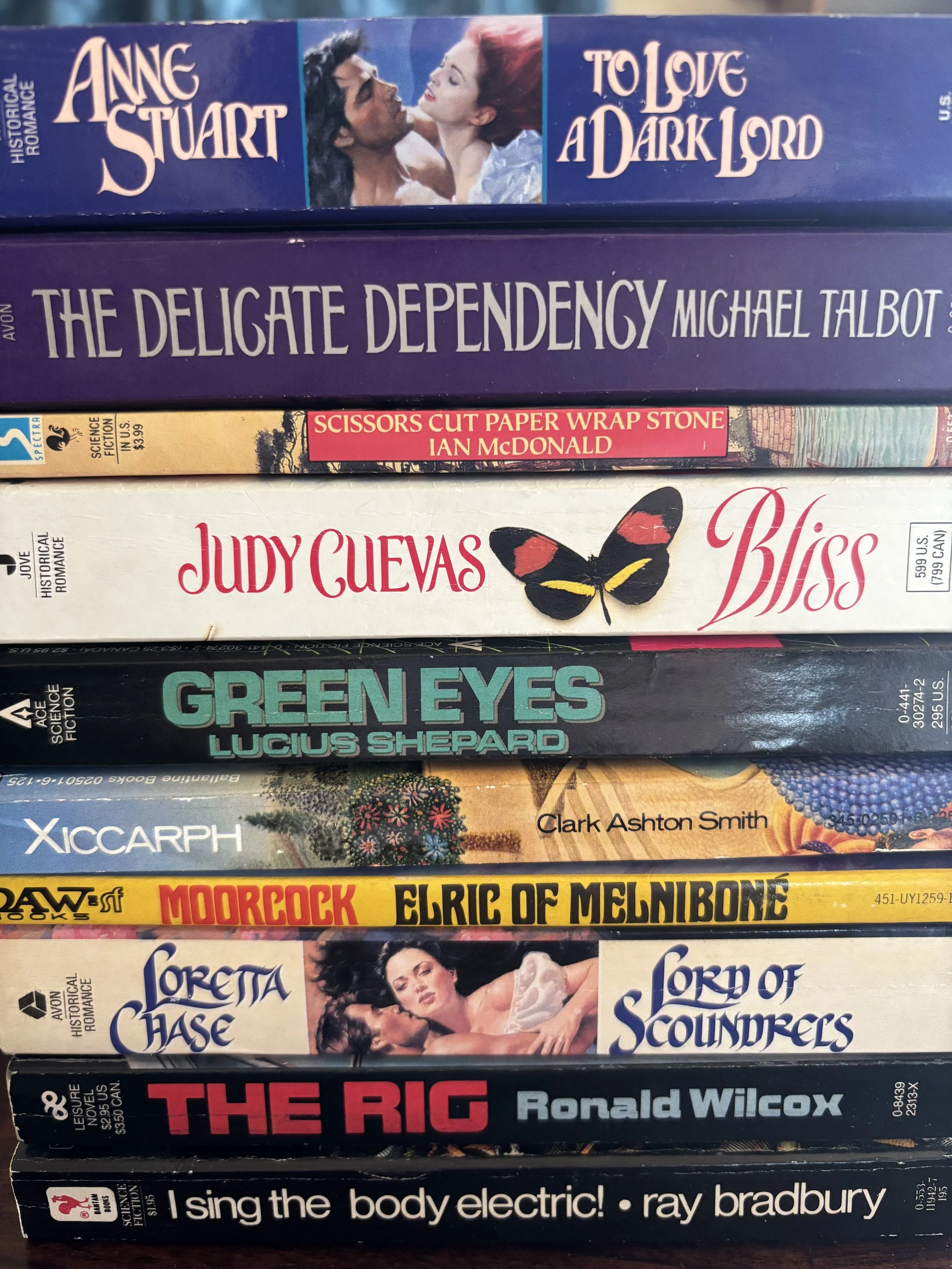 Stack of seven paperback books with colorful covers, featuring titles such as "To Love A Dark Lord," "The Delicate Dependency," "Scissors Cut Paper Wrap Stone," "Bliss," "Green Eyes," "Sorte di Chase," and "I Sing the Body Electric!" by Ray Bradbury.
