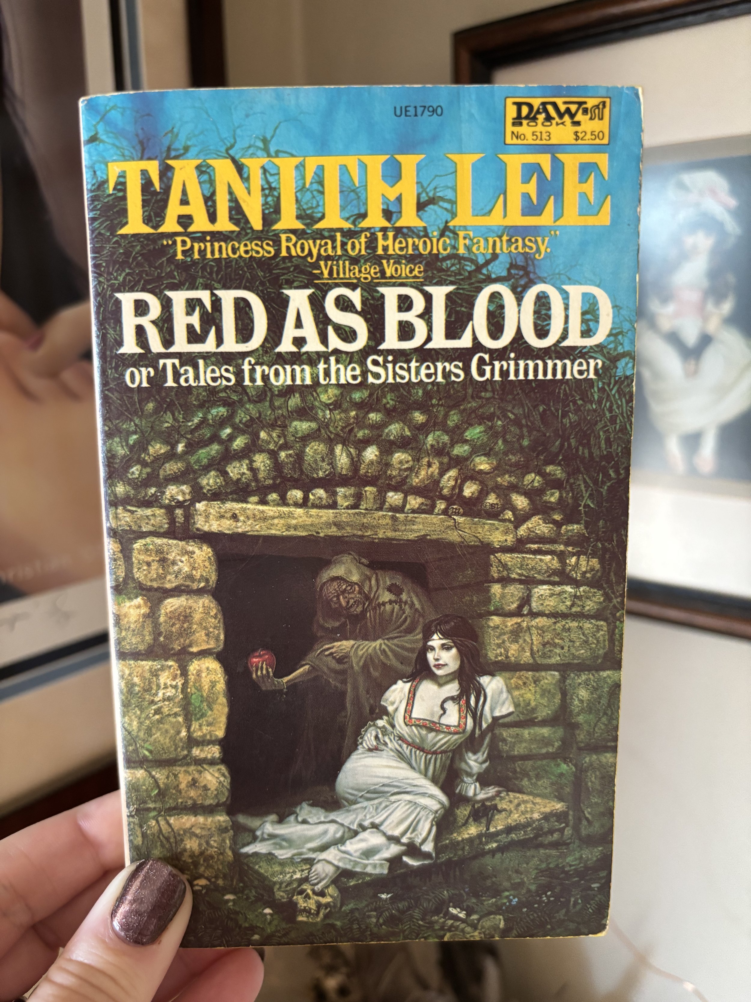 A book titled "Red as Blood or Tales from the Sisters Grimmer" by Tanith Lee being held up, with a fantasy style cover illustration of a woman in a white dress sitting beside a devil-like figure with horns and long hair, with a stone archway and foliage in the background.