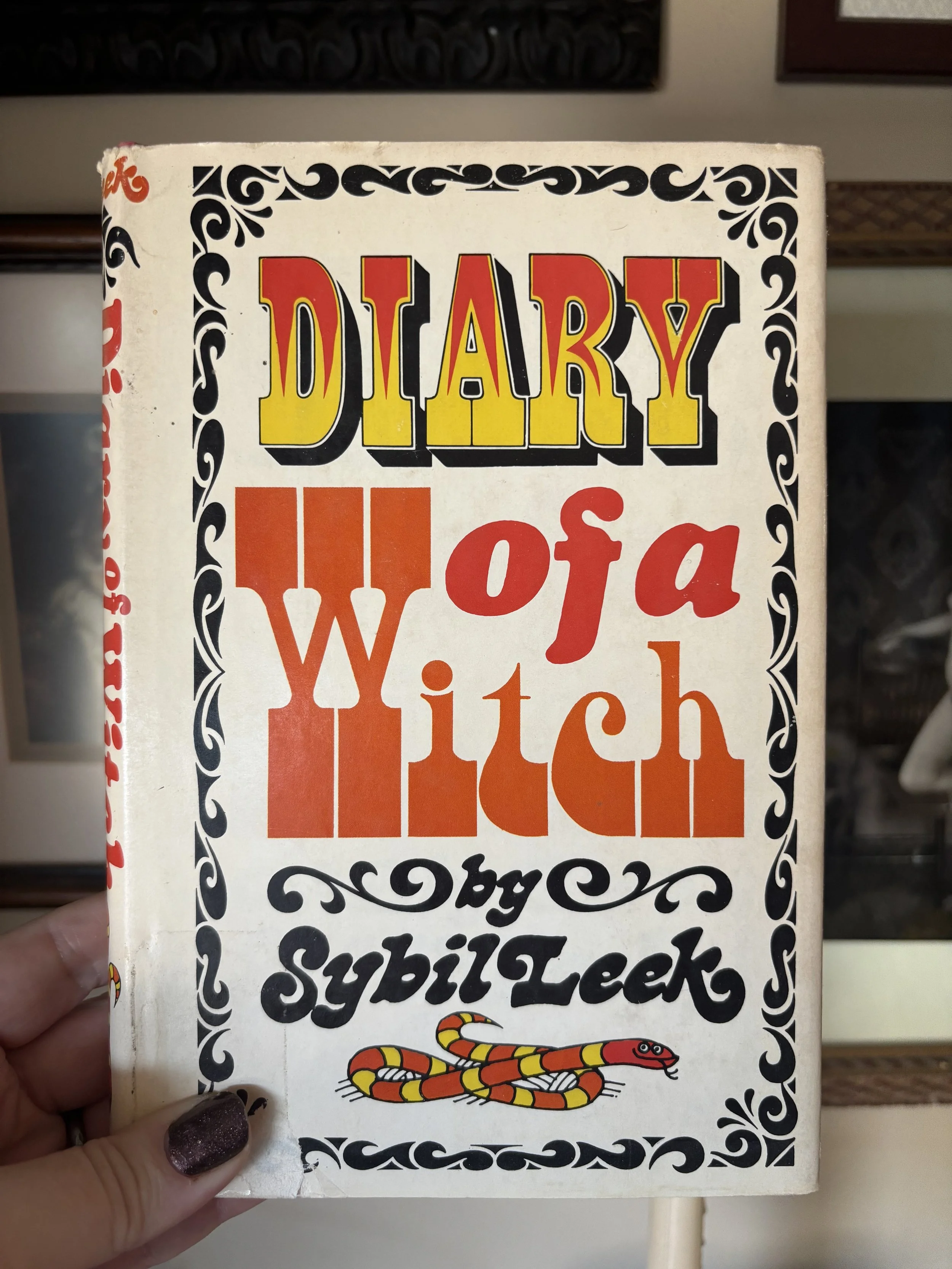 A woman holding a vintage-style paperback book with the title 'Diary of a WITCH' and an illustration of a colorful snake on a white background, with decorative black borders.