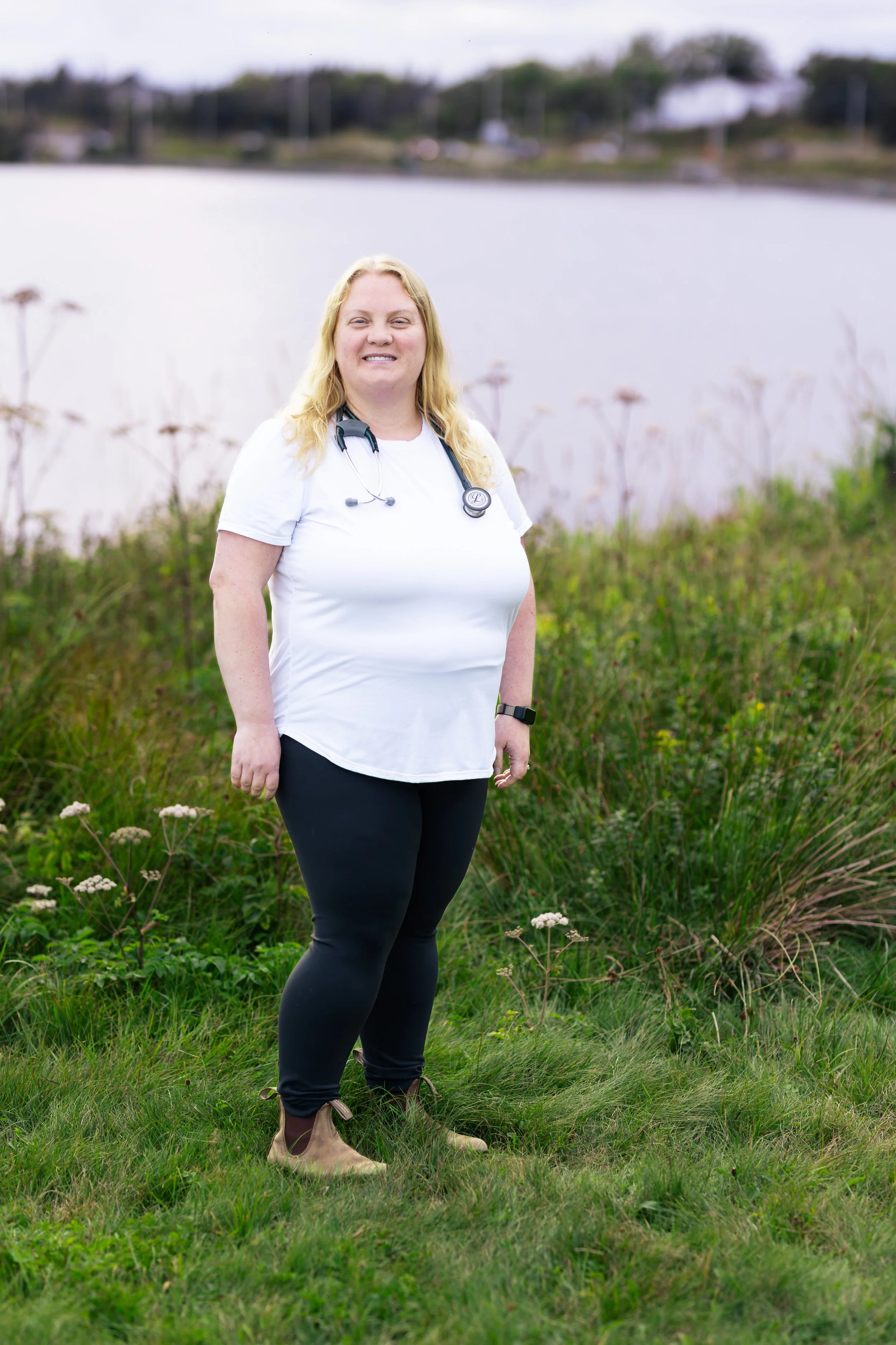 A woman with blonde hair wearing a white shirt with a stethoscope and black pants stands in grass near a body of water with trees and buildings in the background.