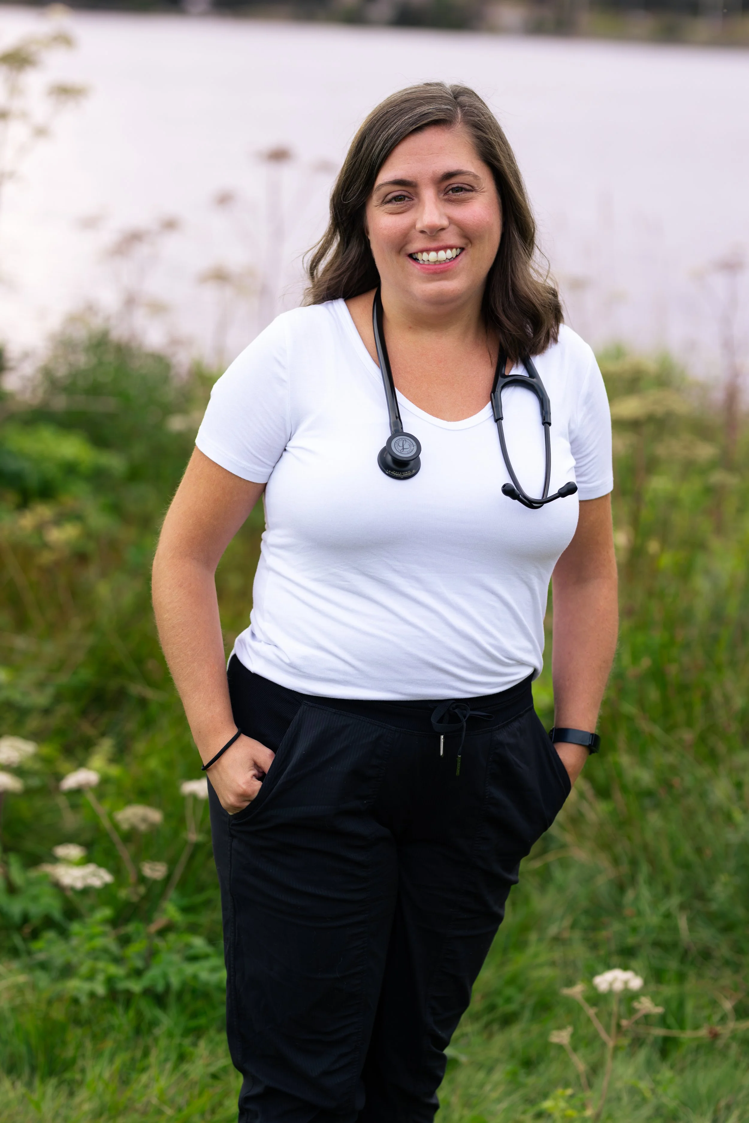 A woman with shoulder-length brown hair and a stethoscope around her neck stands outdoors in a grassy area with blurred plants in the background. She is smiling and wearing a white T-shirt and black pants.