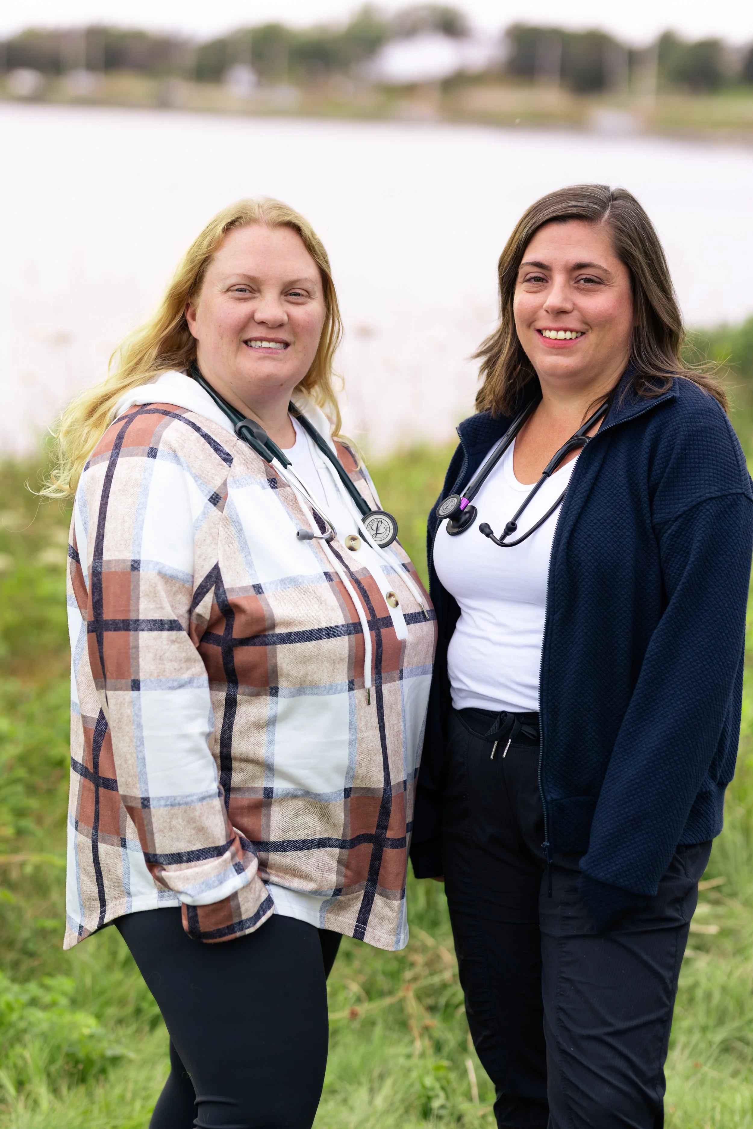 Two female healthcare professionals standing outdoors near a body of water, smiling at the camera, wearing stethoscopes around their necks.