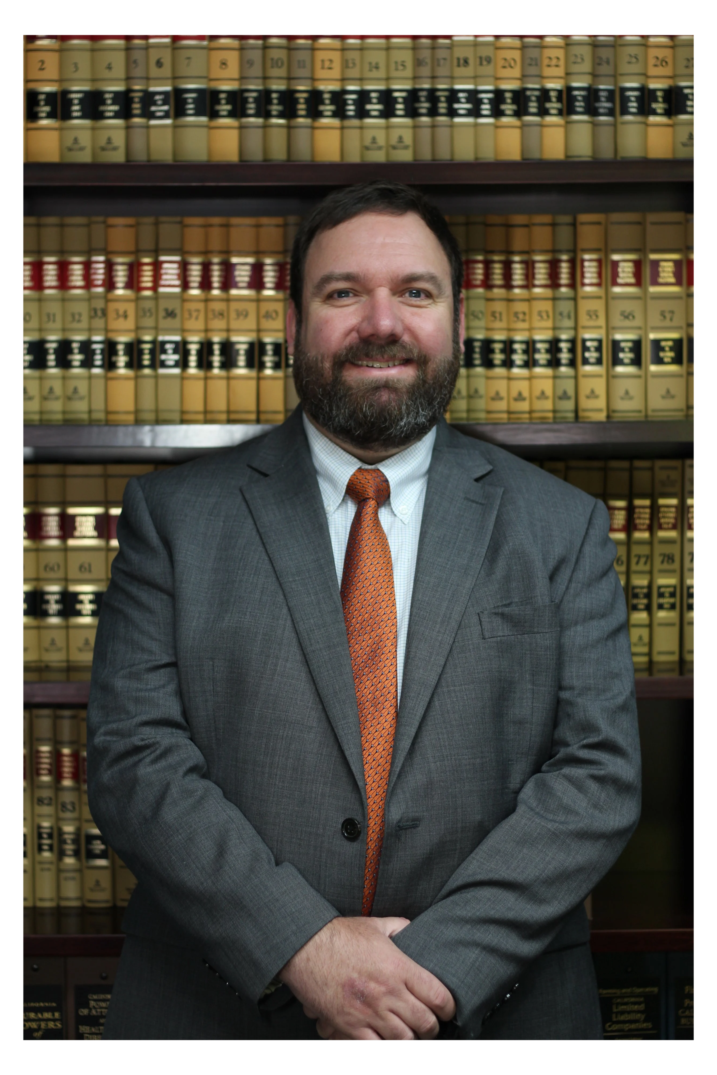 A man with dark hair, a beard, and a mustache wearing a grey suit and orange tie, smiling with arms crossed in front of bookshelves filled with legal books.