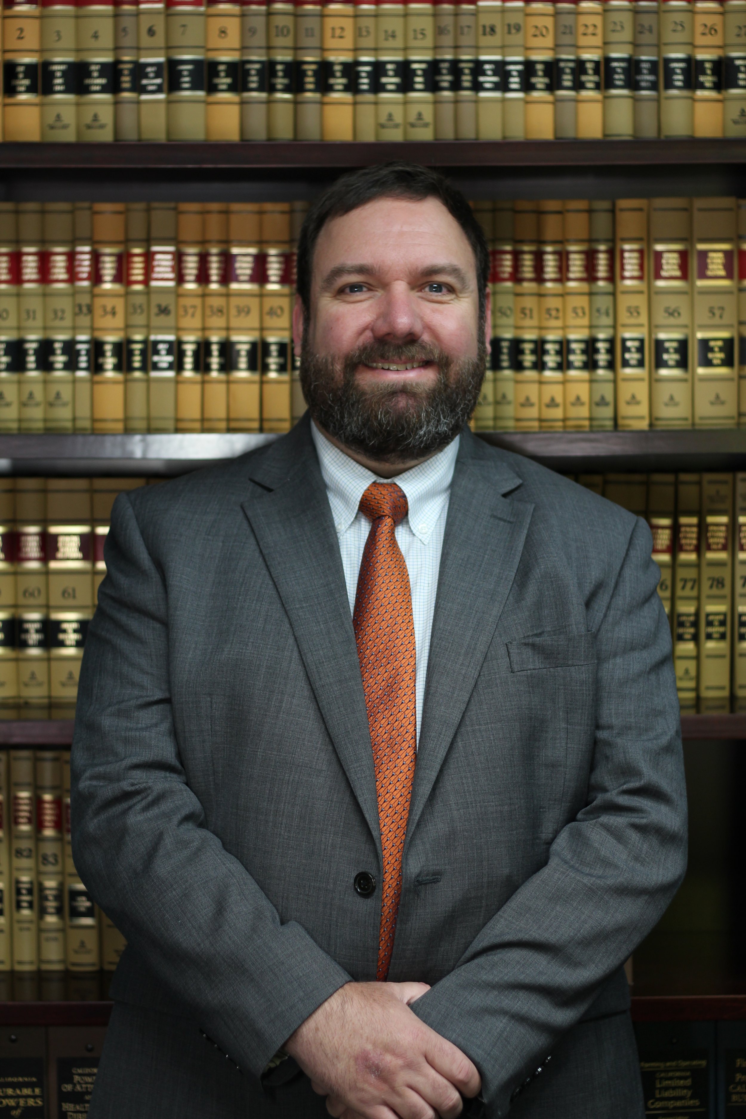 A man with a beard wearing a gray suit and an orange tie standing in front of a bookshelf filled with legal books.
