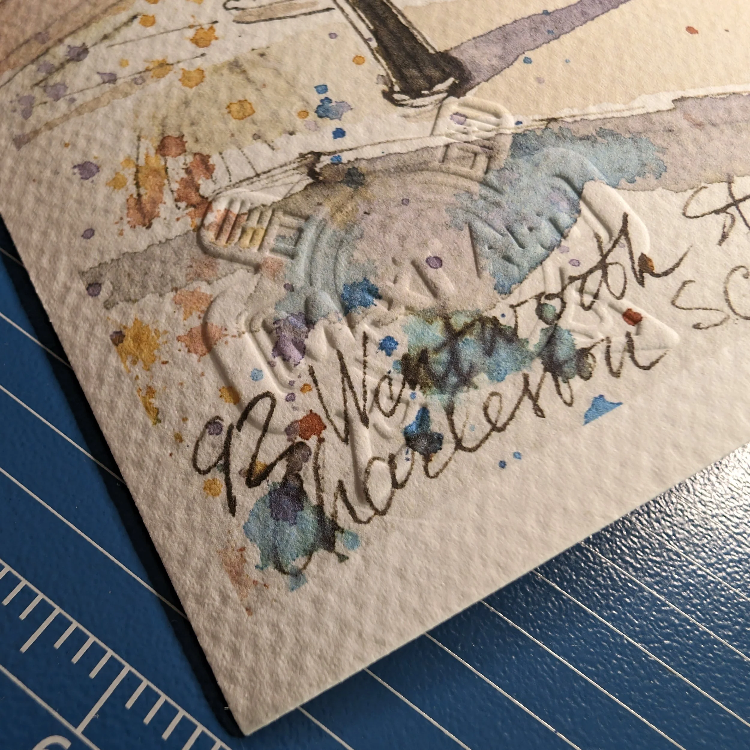 Close-up of a high quality cotton textured paper with handwritten text and watercolor splashes, featuring MaxT Art logo imprint and decorative elements in the background.