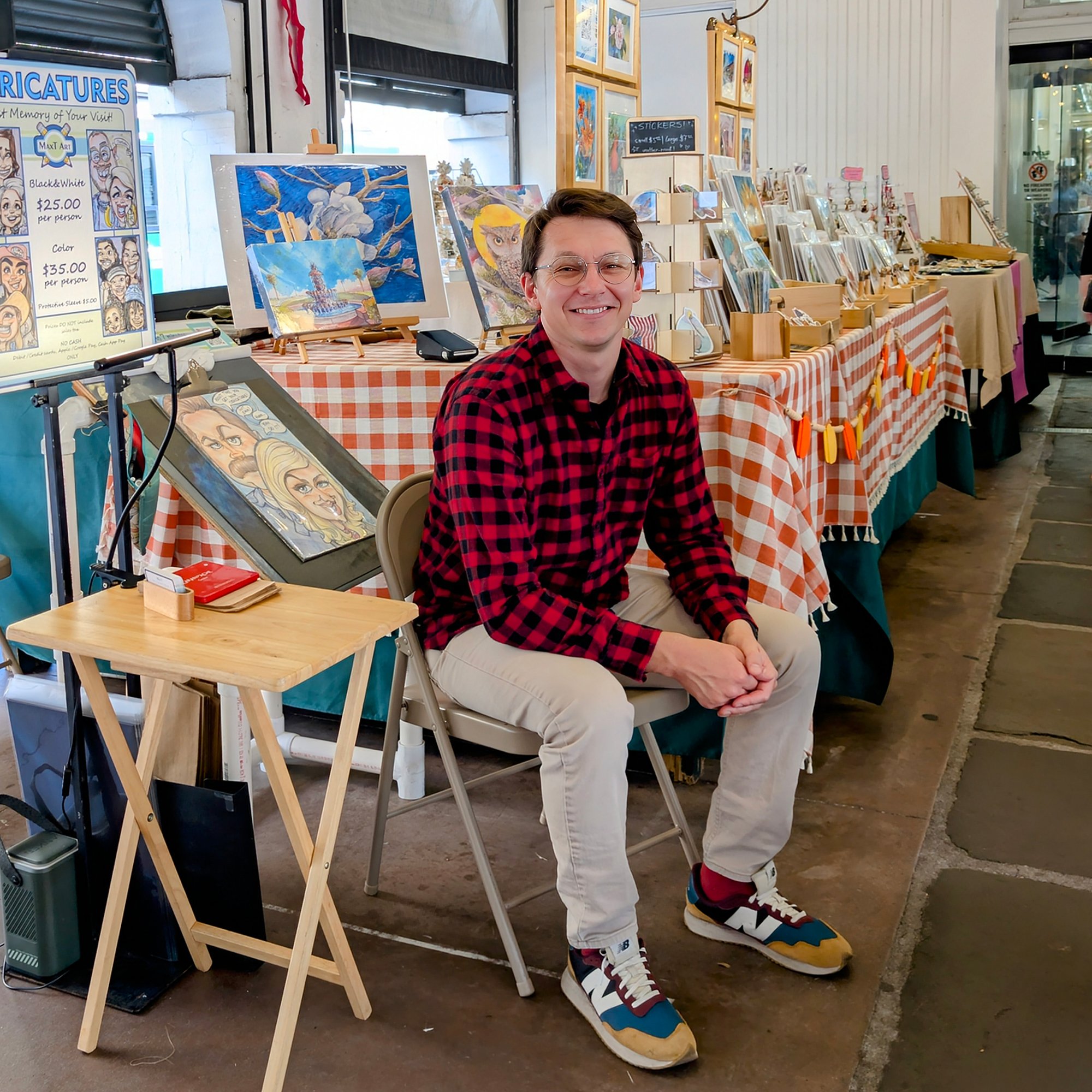 Max is sitting in front of his art booth at the Charleston City Market. The booth displays Max's Charleston, Bird, and Flower paintings,. The background features a sign advertising caricatures, along with framed artwork hanging on the wall
