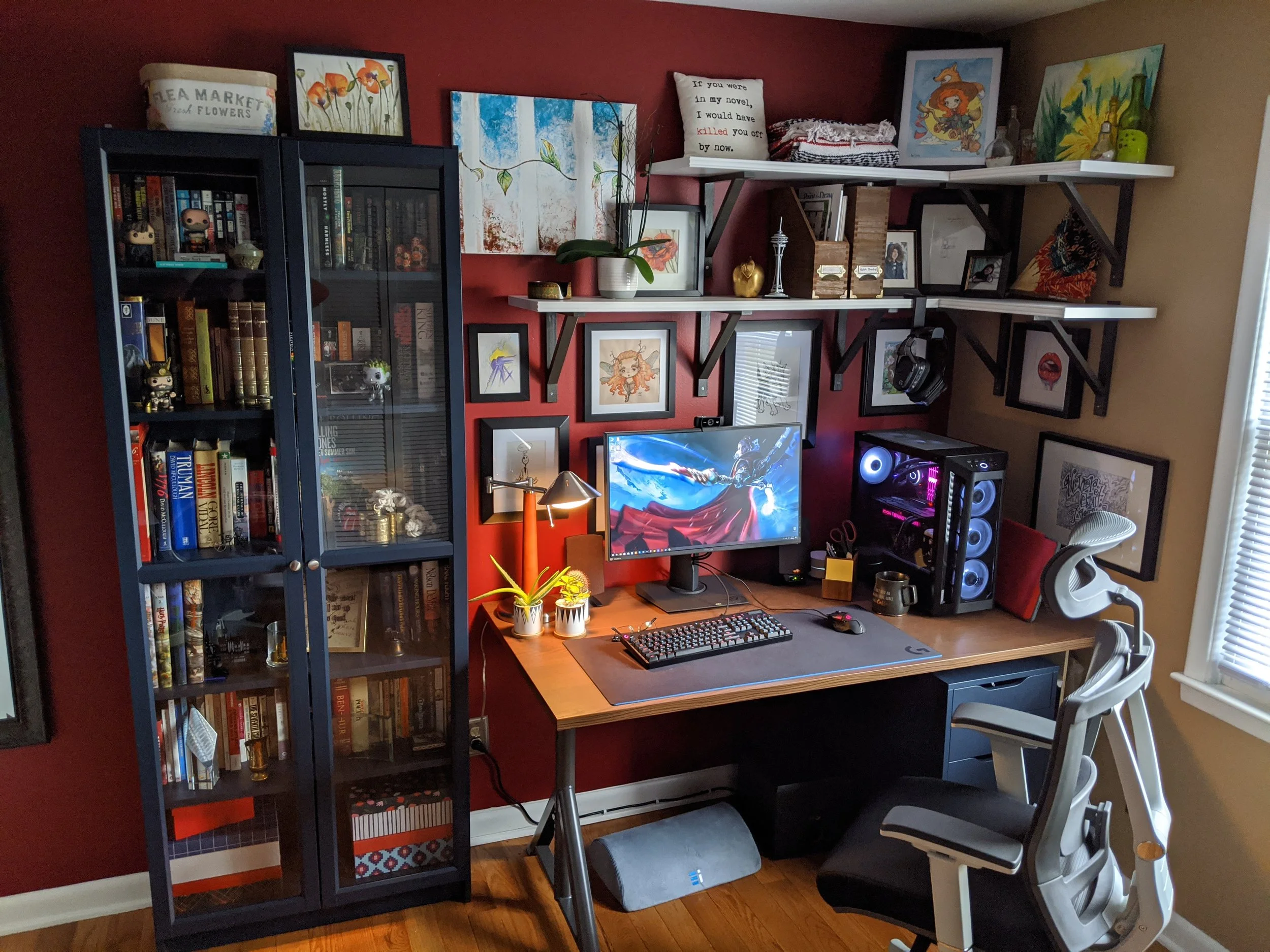 A home office with a wooden desk, computer monitor, black computer tower with RGB lighting, white office chair, black bookshelf filled with books and figures, wall art including paintings and framed pictures, plants, and various decorations arranged on shelves and tables.