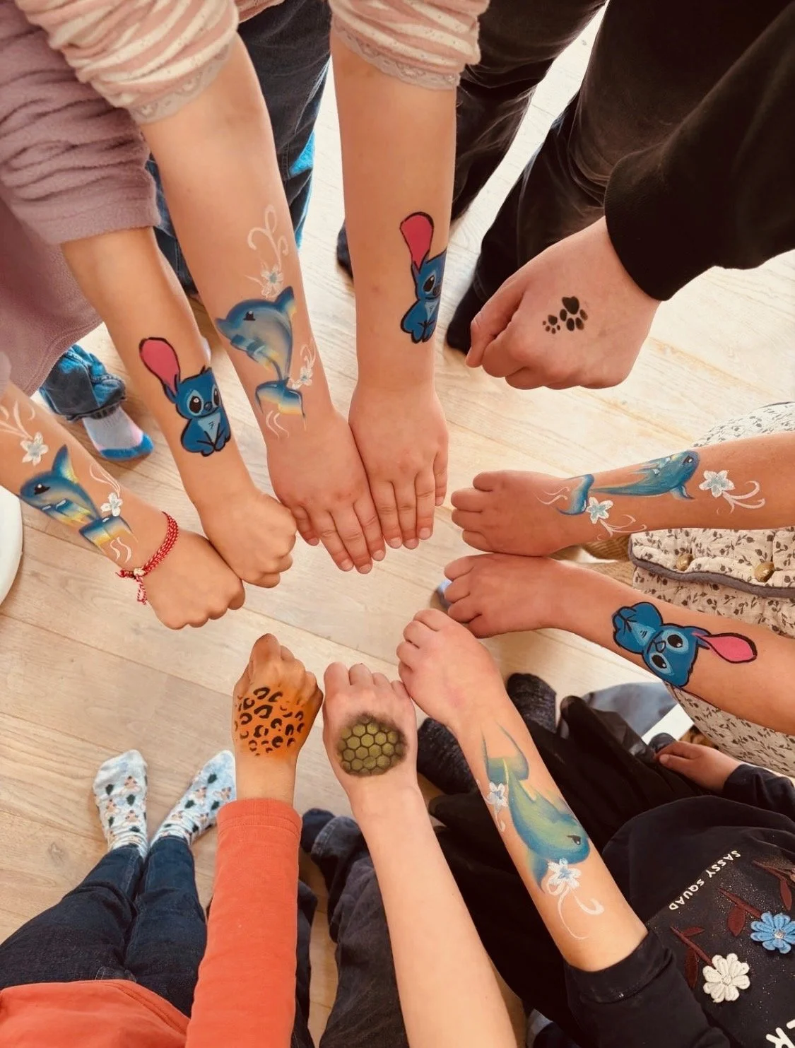 Mehrere Kinder und Erwachsene zeigen ihre Armen mit geschminkten Motiven , darunter Delfin , Stich, Tiere und andere kreative Designs.