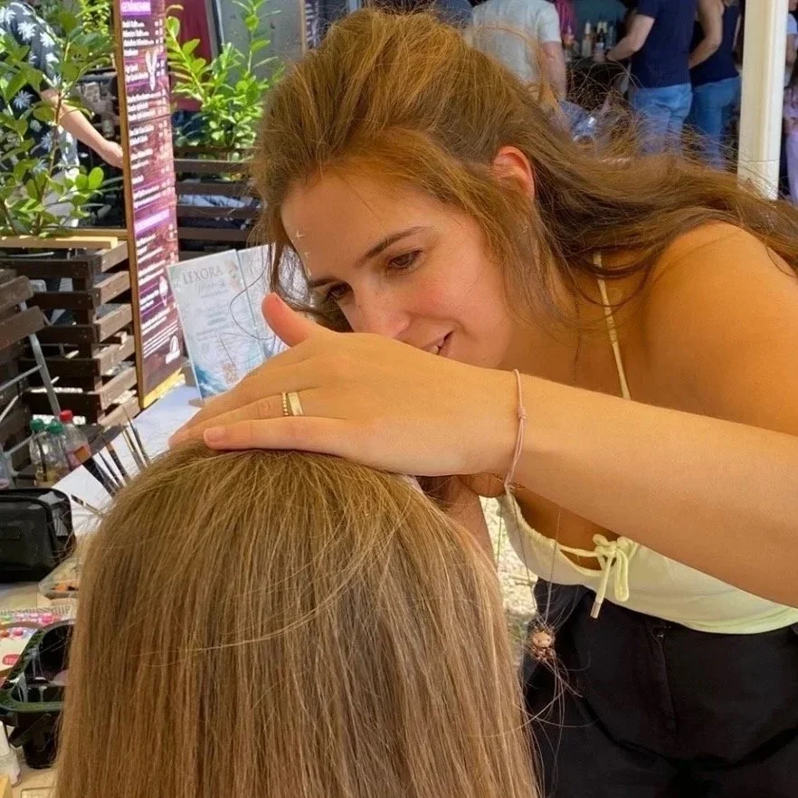 Frau beim Schminken einer anderen Person. Sie hat lange Haare, trägt ein weißes Oberteil und einen Haarreif. Im Hintergrund sind weitere Menschen und eine Pappwand mit Pflanzen.